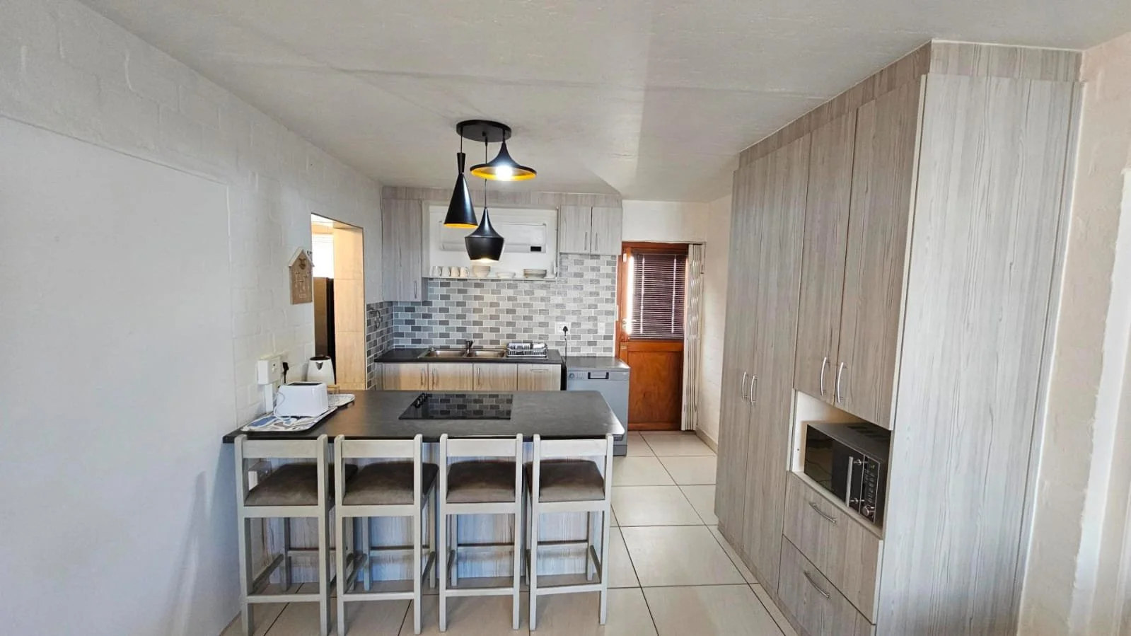Kitchen with light wood cabinets, a black countertop island with four white chairs, gray tiled backsplash, a small window with brown blinds, stainless steel sink, white stove, microwave, and modern black pendant lights.