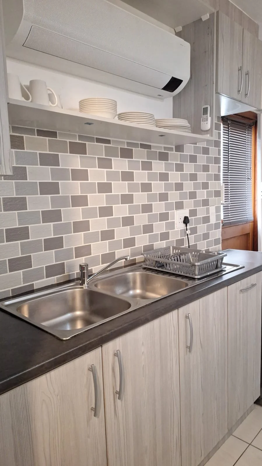 Kitchen with double sink, gray mosaic tile backsplash, open shelves with white dishes, and wooden cabinets.