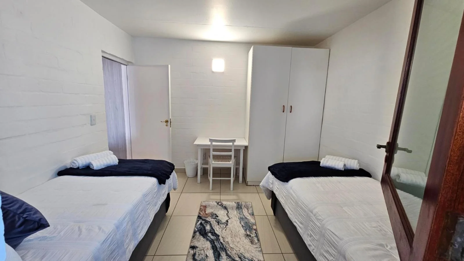A small bedroom with two twin beds, black and white bedding, towels, a rug, a white wardrobe, a small table with chairs, and a mirror.