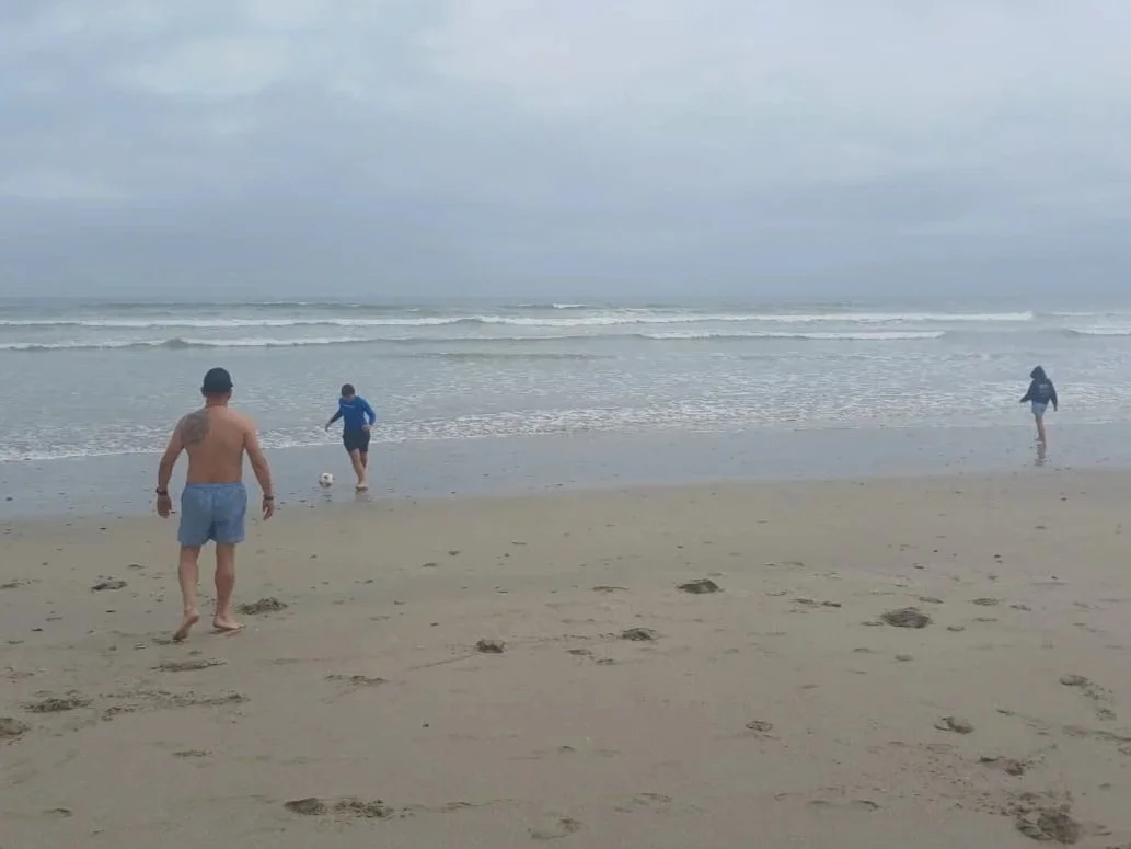 People playing soccer on a cloudy beach with footprints in the sand.