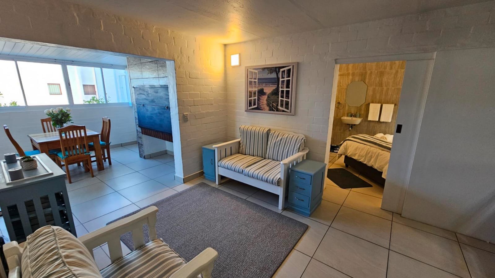 Living room with white brick walls, blue and white striped furniture, adjacent dining area with wooden table and chairs, and a view into a bedroom with a bed and desk