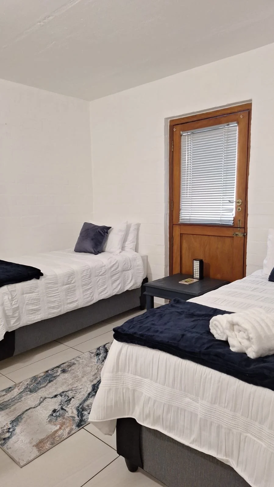 A bedroom with two twin beds with white bedspreads and dark pillows, a black nightstand with a remote control on top, a small patterned rug on the tiled floor, and a wooden door with blinds on the window
