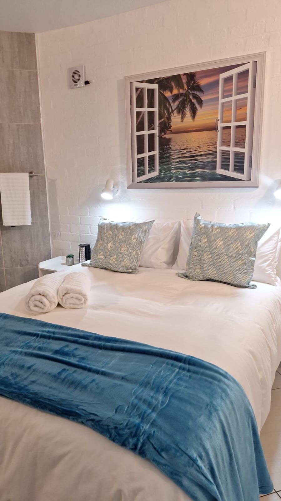 Hotel room with a bed, decorative throw pillows, rolled towels, a window with a sunset over the ocean, and tropical scenery.