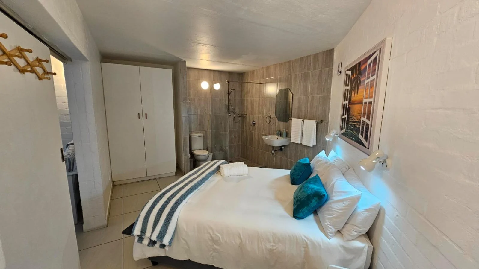 Hotel room with a bed, blue pillows, and a tropical sunset photo on the wall; open closet, small bathroom with shower, toilet, and sink.