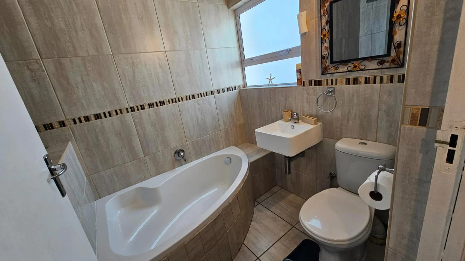Small bathroom with a bathtub, a wall-mounted sink, a toilet, a window with a starfish decoration, a mirror with a floral frame, and beige tiled walls.
