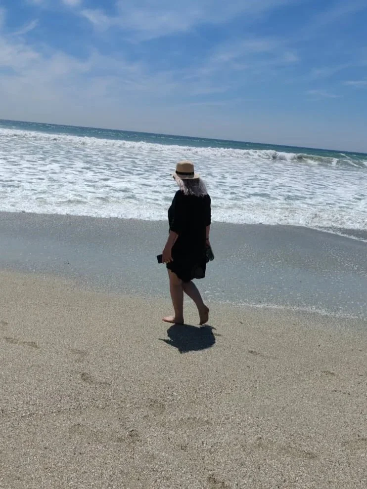 A woman walking on a sandy beach near the ocean, wearing a black dress and a sunhat, holding something in her right hand, with the sky partly cloudy.