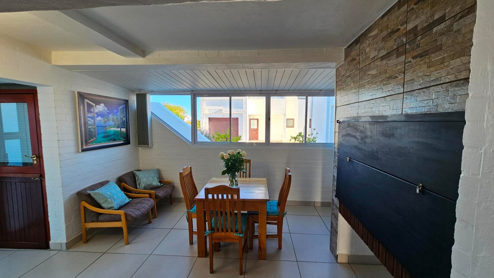 A cozy dining area with a wooden table surrounded by four chairs, a small sofa with pillows, and a vase of flowers on the table, with large windows providing natural light and a view of neighboring buildings, in a room with white brick walls, tiled f