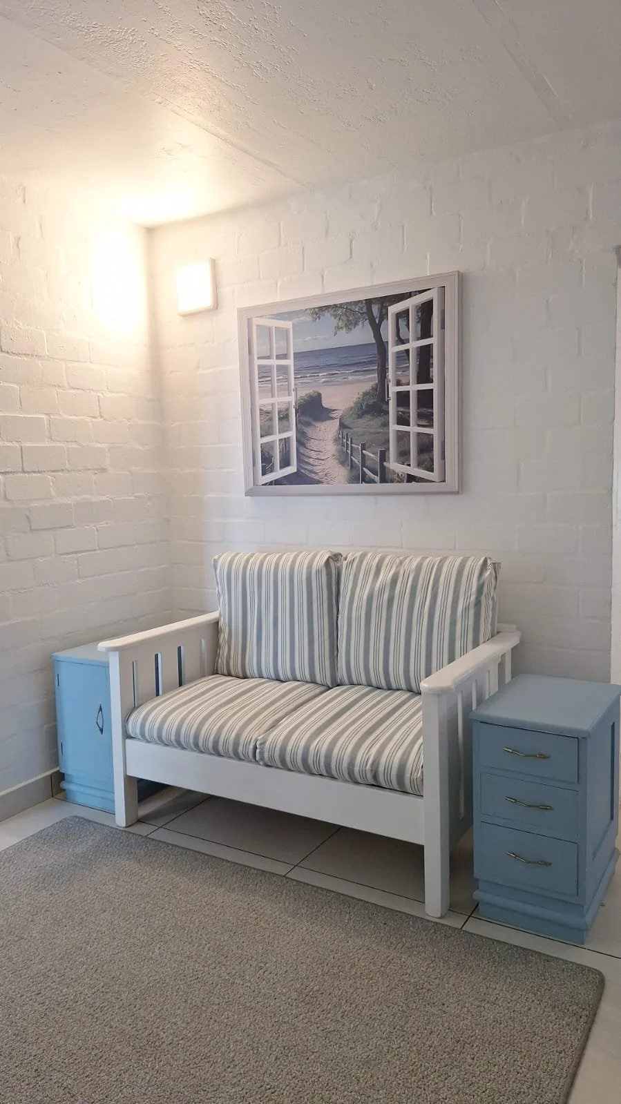 A cozy room corner with a white wooden bench that has striped cushions, a blue side table with three drawers, and a framed picture of an open window overlooking a beach scene on the wall.