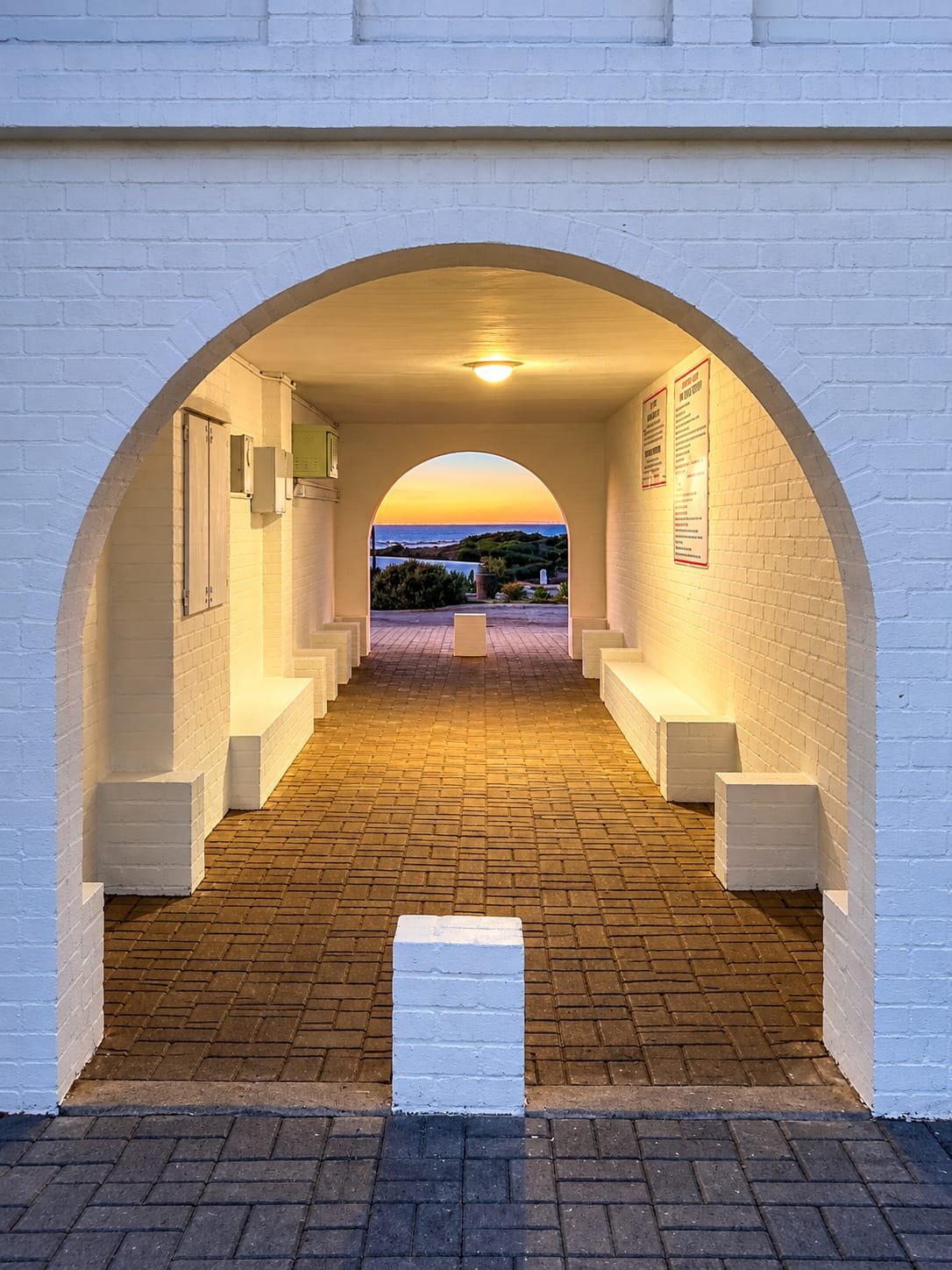 View through a brick archway outdoor corridor at sunset, with white brick walls, a brick floor, and benches on the sides, overlooking a scenic landscape with greenery and water.