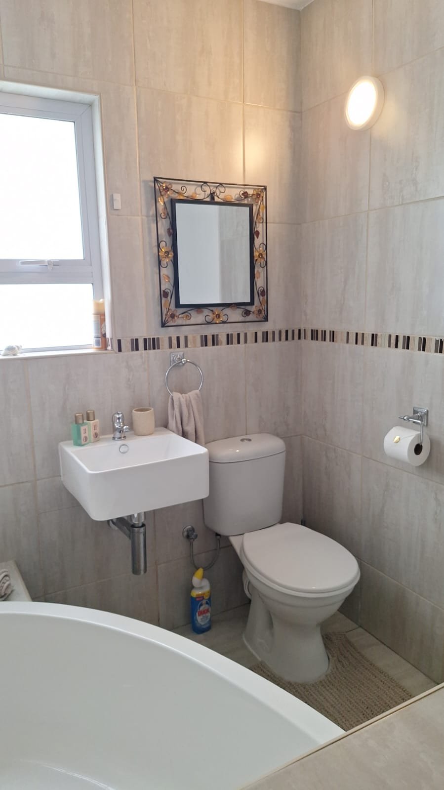 A small bathroom with beige tiled walls, a window, a small white sink, a toilet with a closed lid, a decorative mirror, a hand towel, toilet paper, and cleaning supplies.
