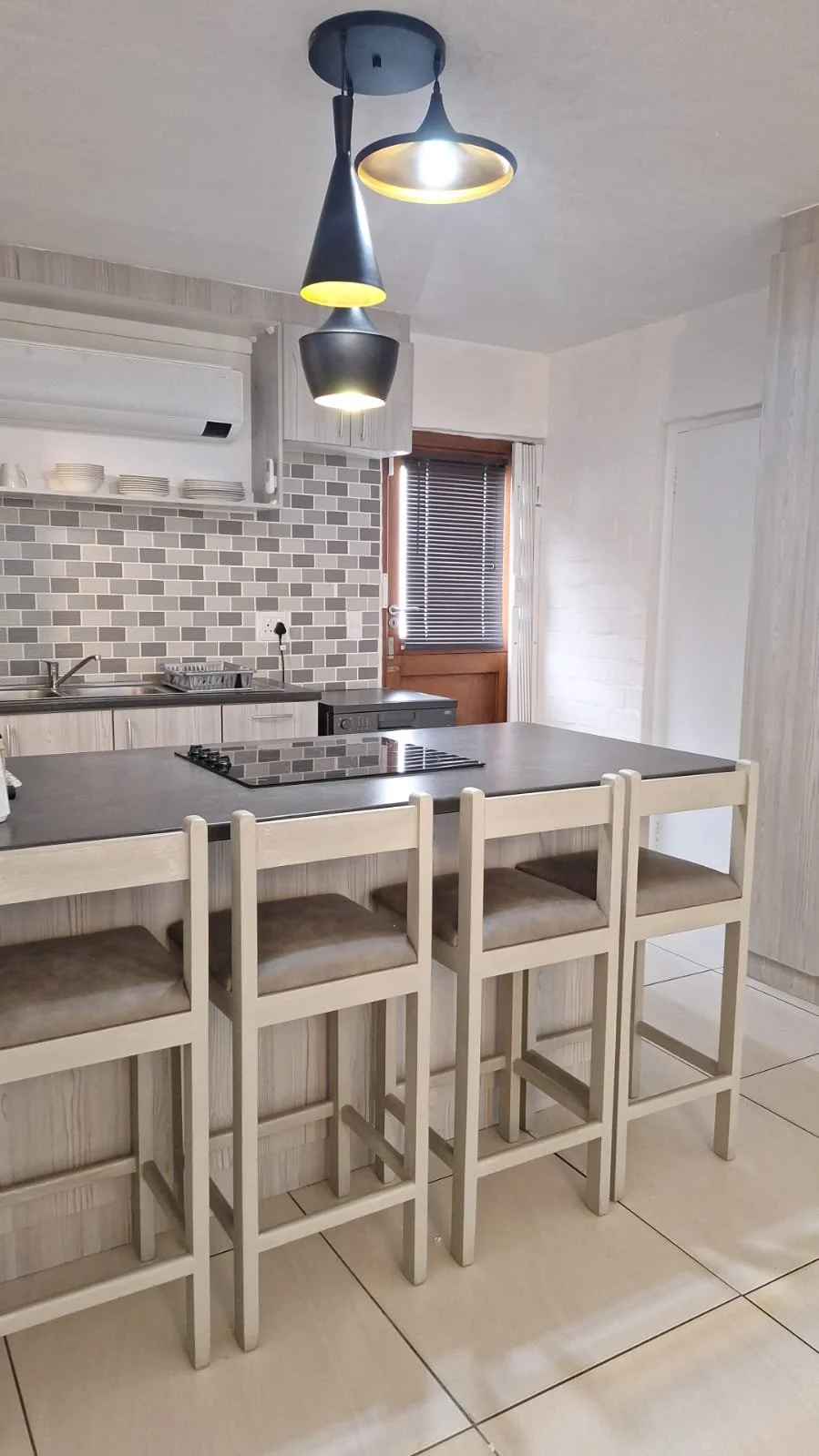 Modern kitchen with four beige barstools at a gray breakfast bar, black stovetop, gray tiled backsplash, white cabinets, window with blinds, and a black hanging ceiling light.