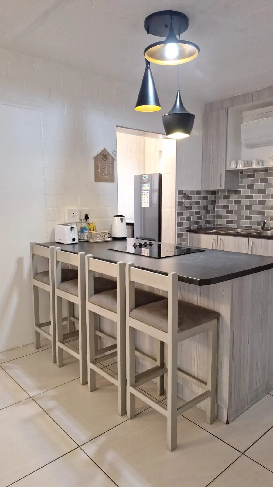 A modern kitchen with a breakfast bar and four white chairs, featuring a pendant lighting fixture hanging from the ceiling, a fridge, a stove, and a tiled backsplash.