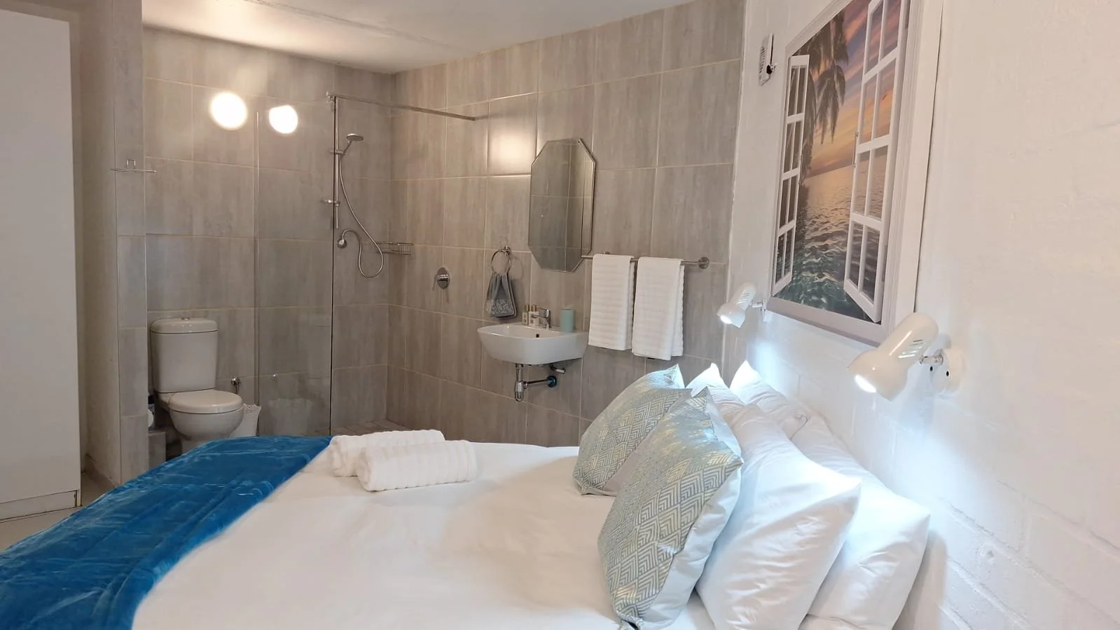 A cozy bedroom with a bed covered in white and blue bedding, featuring pillows and a rolled towel on top. To the right, a wall-mounted picture of a tropical sunset scene with palm trees. Above the bed, adjustable reading lights. In the background, a 