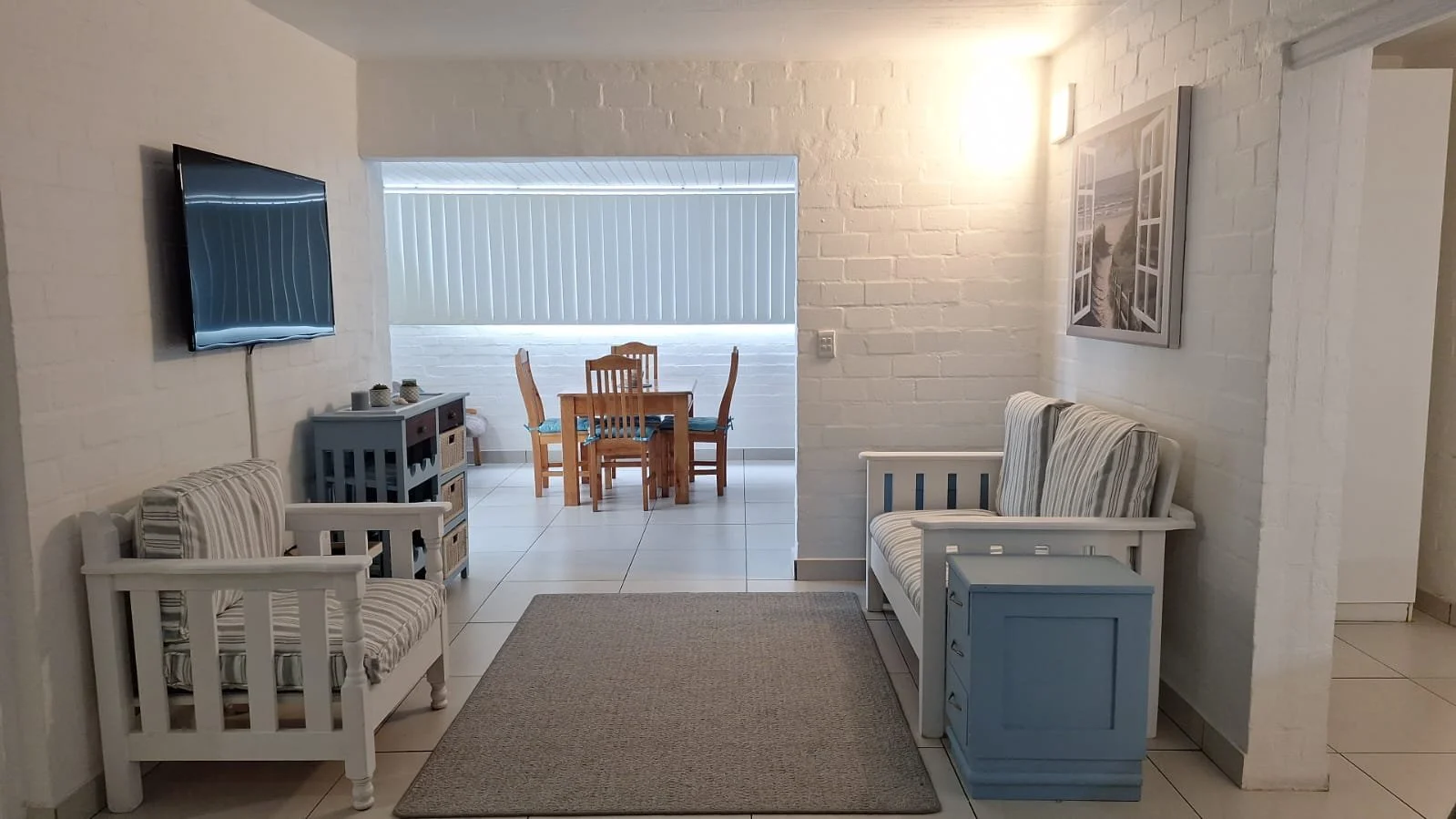 Living room with white brick walls, a wall-mounted flat-screen TV, a white bench with striped cushions, a blue side table, and a picture of a bridge on the wall; in the background, a dining area with a wooden table and chairs.