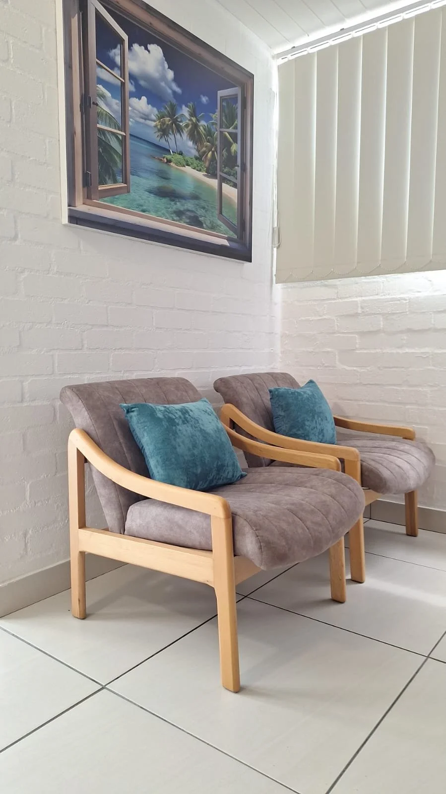 Two wooden-framed chairs with cushioned seats and blue pillows, placed against a white brick wall, with a tropical beach scene window decoration above.
