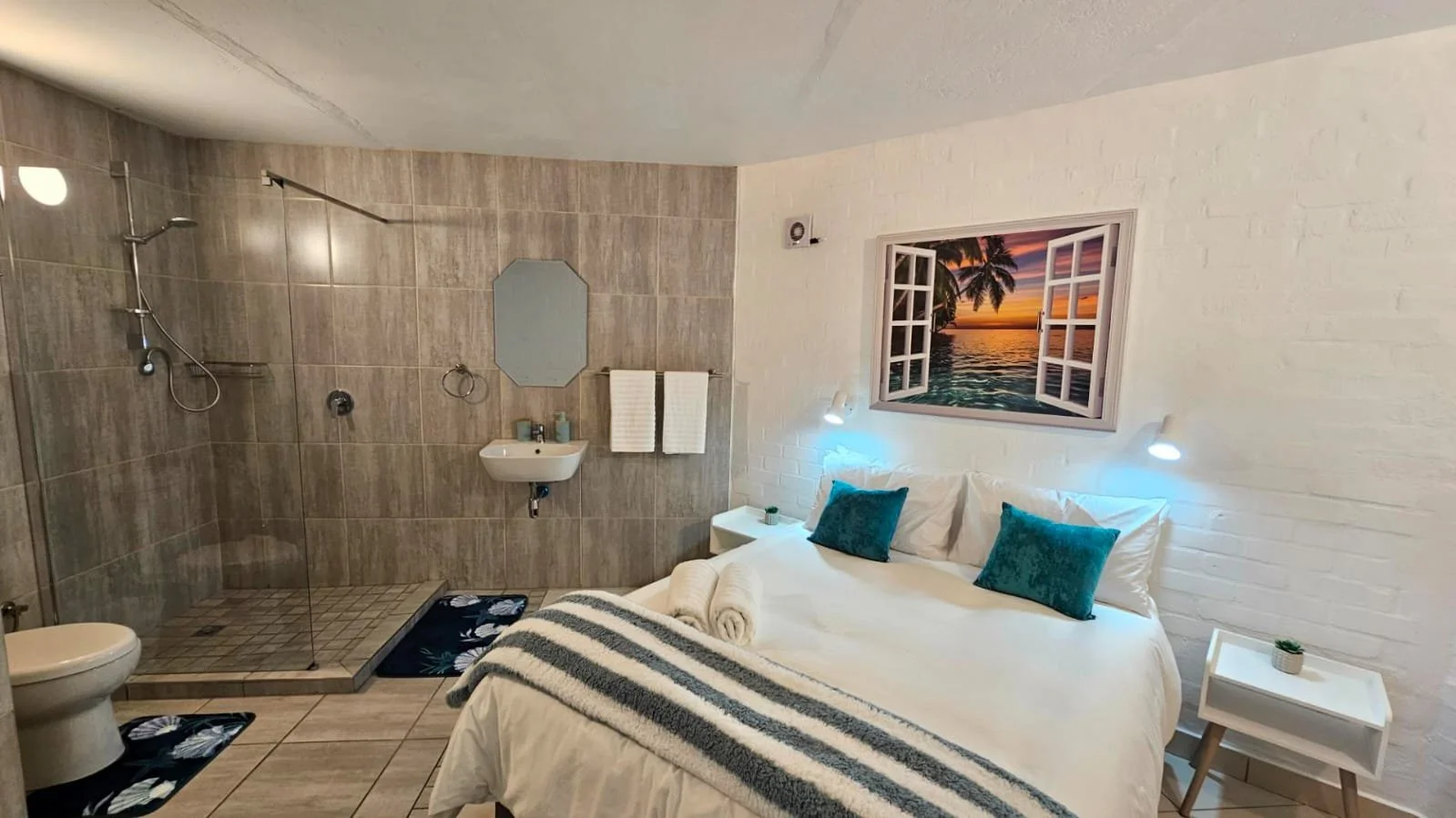 A bedroom with a bed decorated with two teal pillows and a striped blanket, white side tables with a small plant, and a large window view of a tropical sunset with palm trees over water. Adjacent to the bed is a bathroom area with a walk-in shower, a
