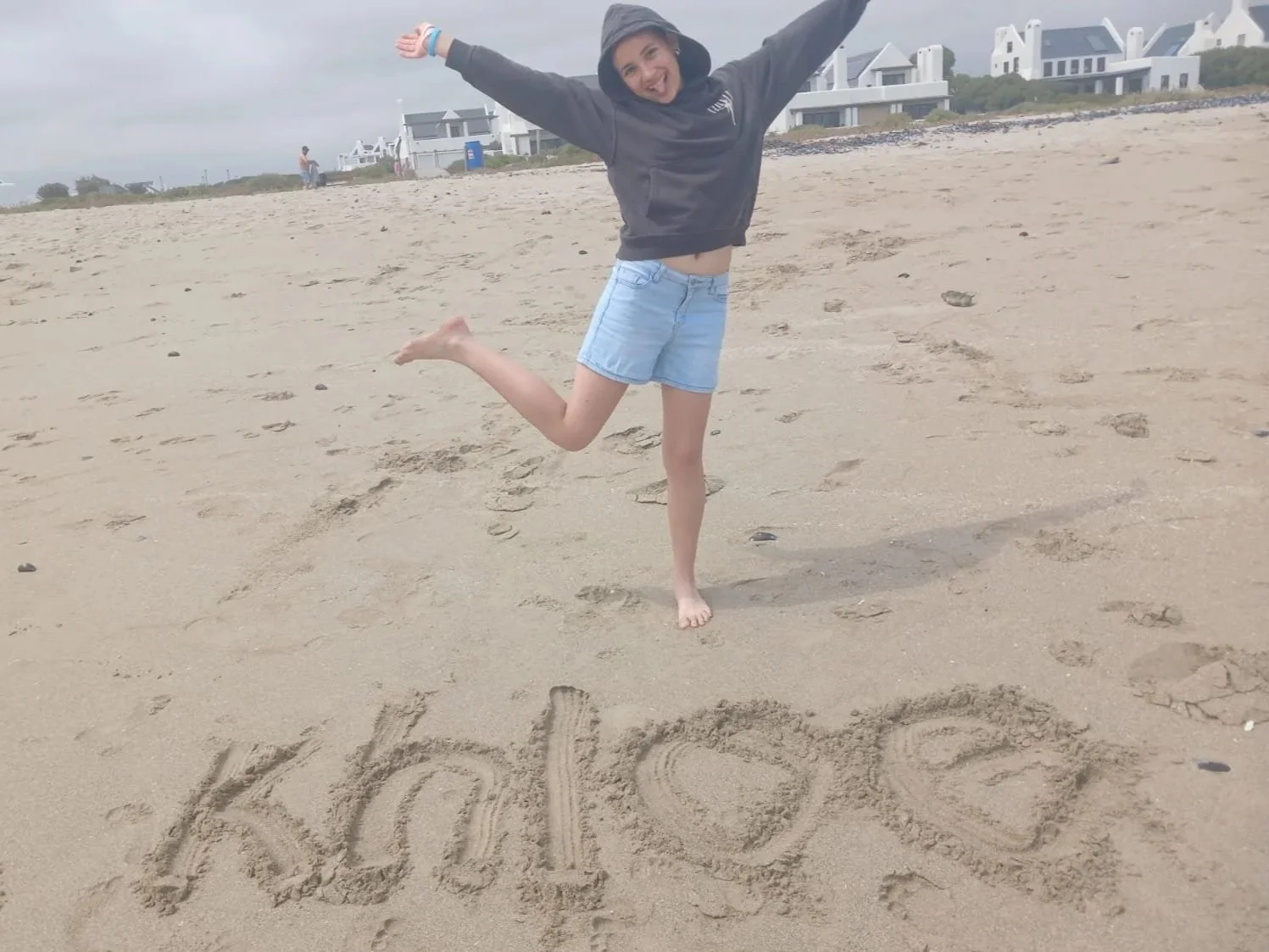 A girl in a black hoodie and denim shorts jumping on a sandy beach with her arms raised. The word 'you' is written in the sand in front of her.