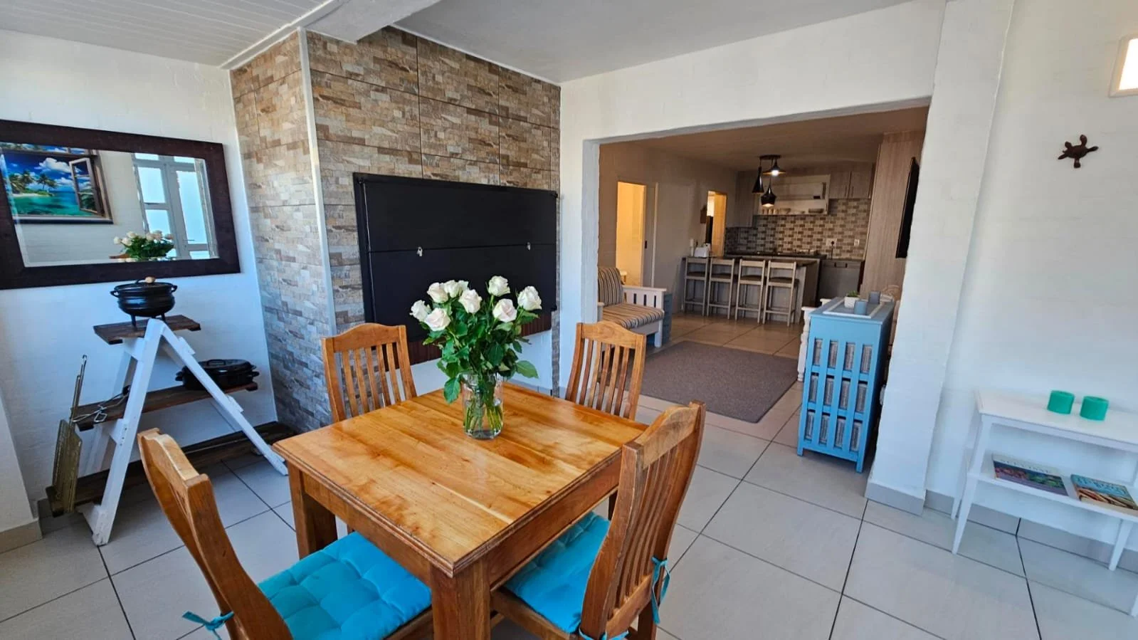 Dining area with a wooden table and chairs, a vase of white roses, and a view into the kitchen with bar stools and a tiled backsplash.