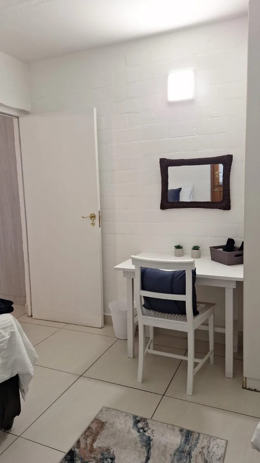 A small white desk with two small potted succulents, a gray basket, and a white chair with a blue pillow, placed against a white brick wall with a rectangular mirror and a wall sconce above.
