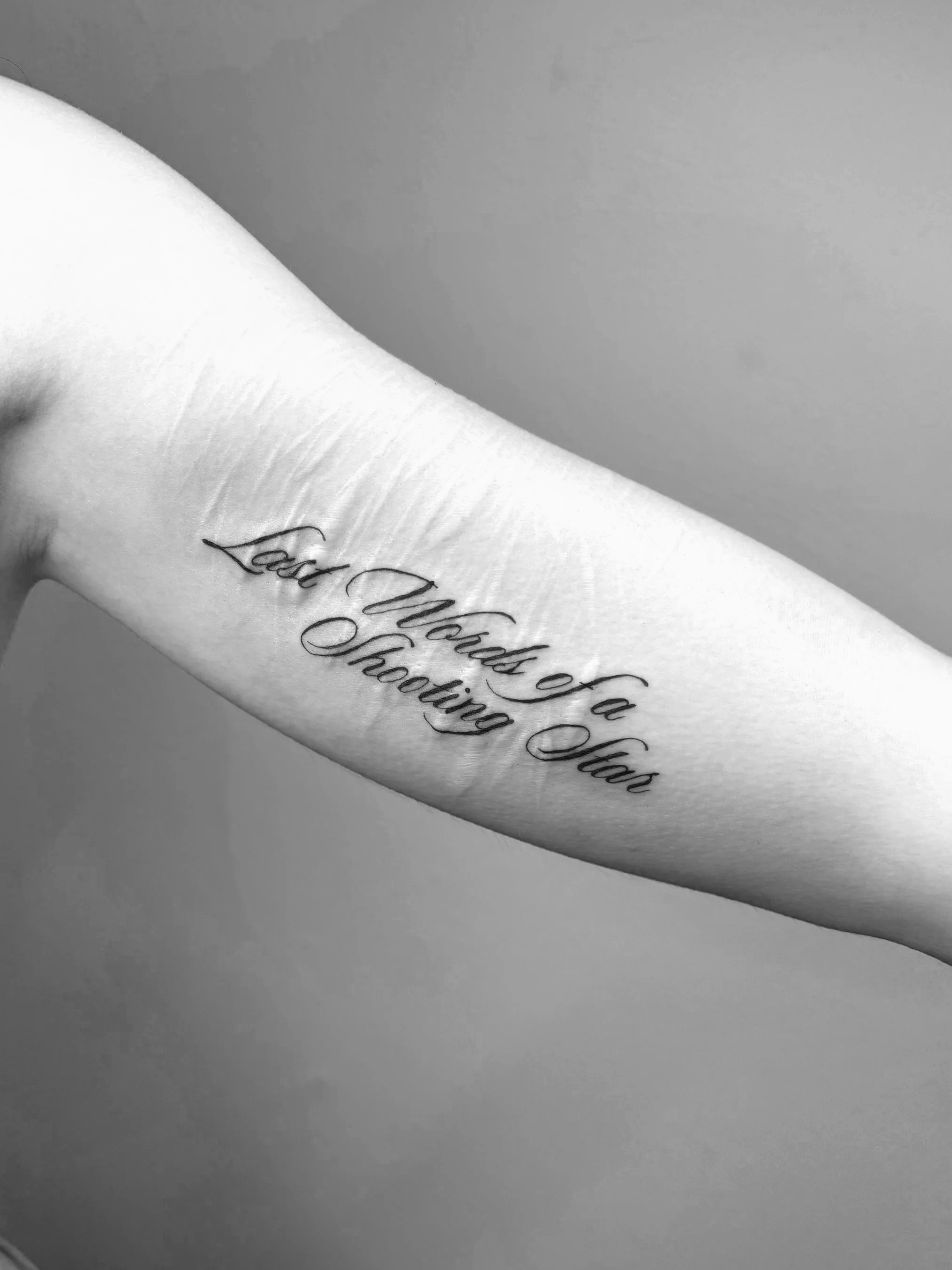 Tattoo auf einem Arm, das die Worte 'Lost Words of a Shooting Star' in kalligraphischer Schrift zeigt.