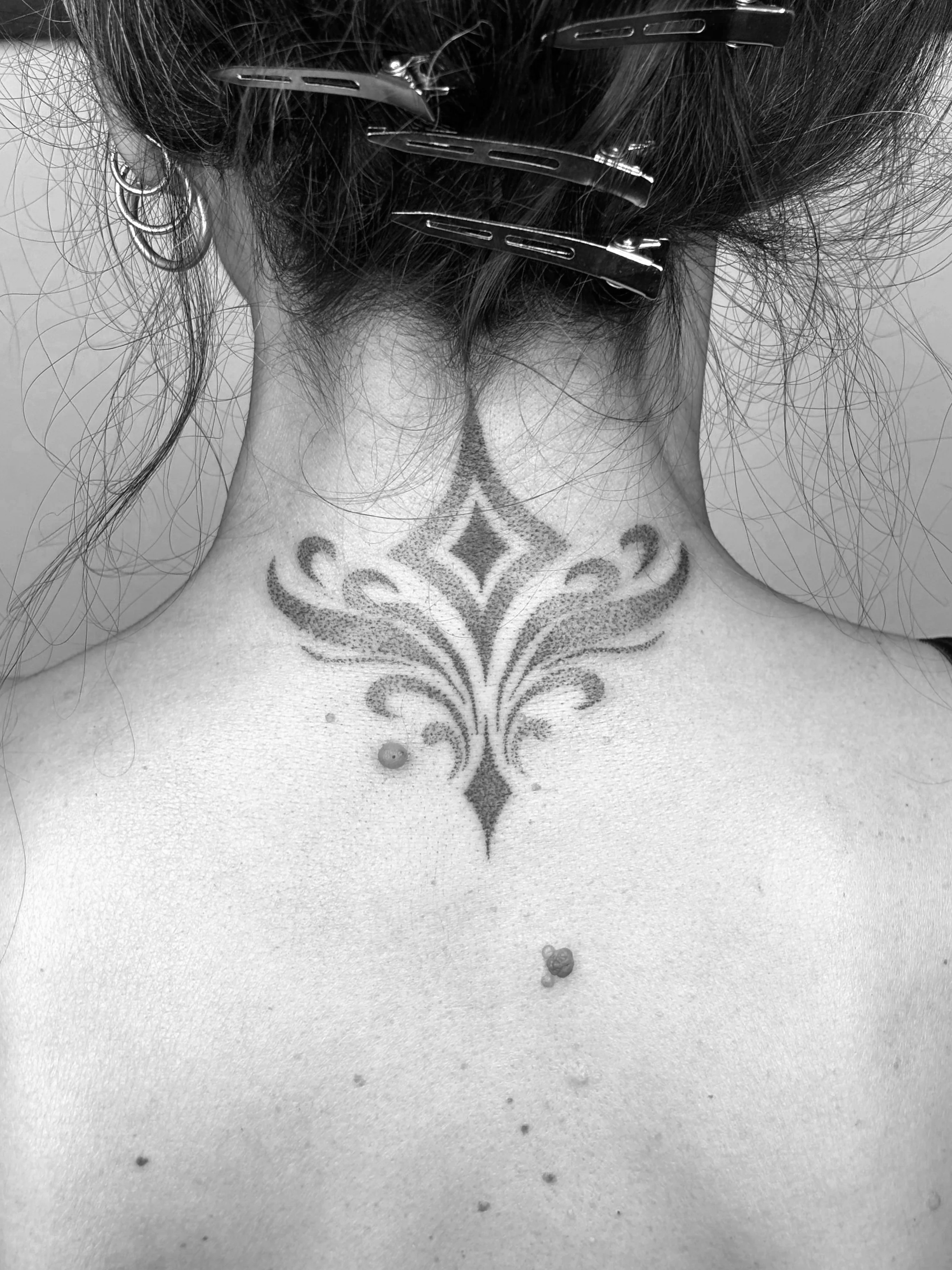 Schwarzes und weißes Foto eines Nacktes Rückens mit Tattoo und mehreren Haarklammern im Haar, Blick auf die Nacken- und Rückenpartie, Zubehör und Piercings sichtbar.