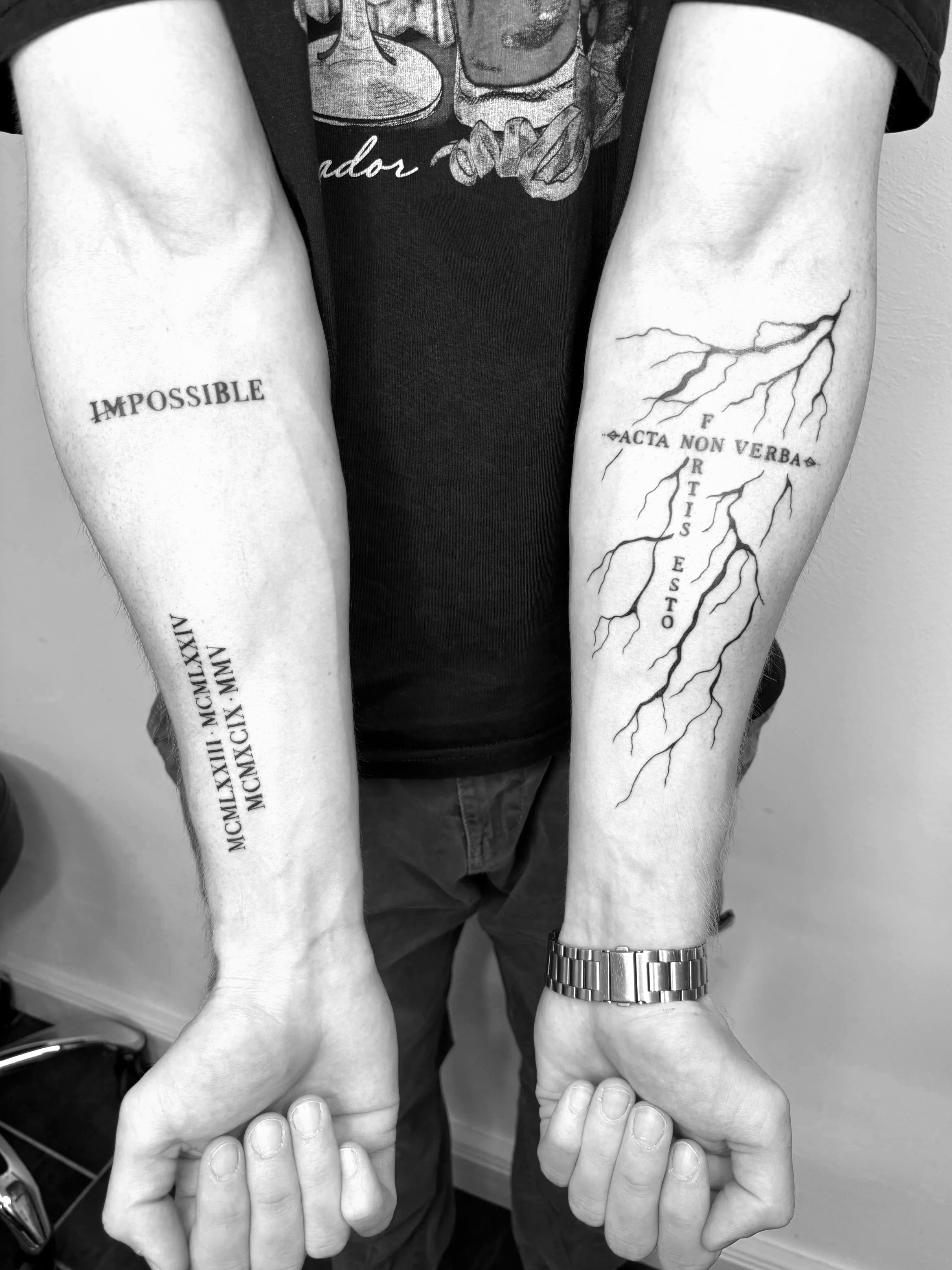 Tattoo auf beiden Armen, das auf der linken Seite das Wort 'UNMÖGLICH' zeigt, und auf der rechten Seite ein Blitz-Design mit lateinischem Spruch 'Acta non verba', zusammen mit Worten 'Justice' und 'Fate' in einer keltischen Schriftart, alle in Schwar