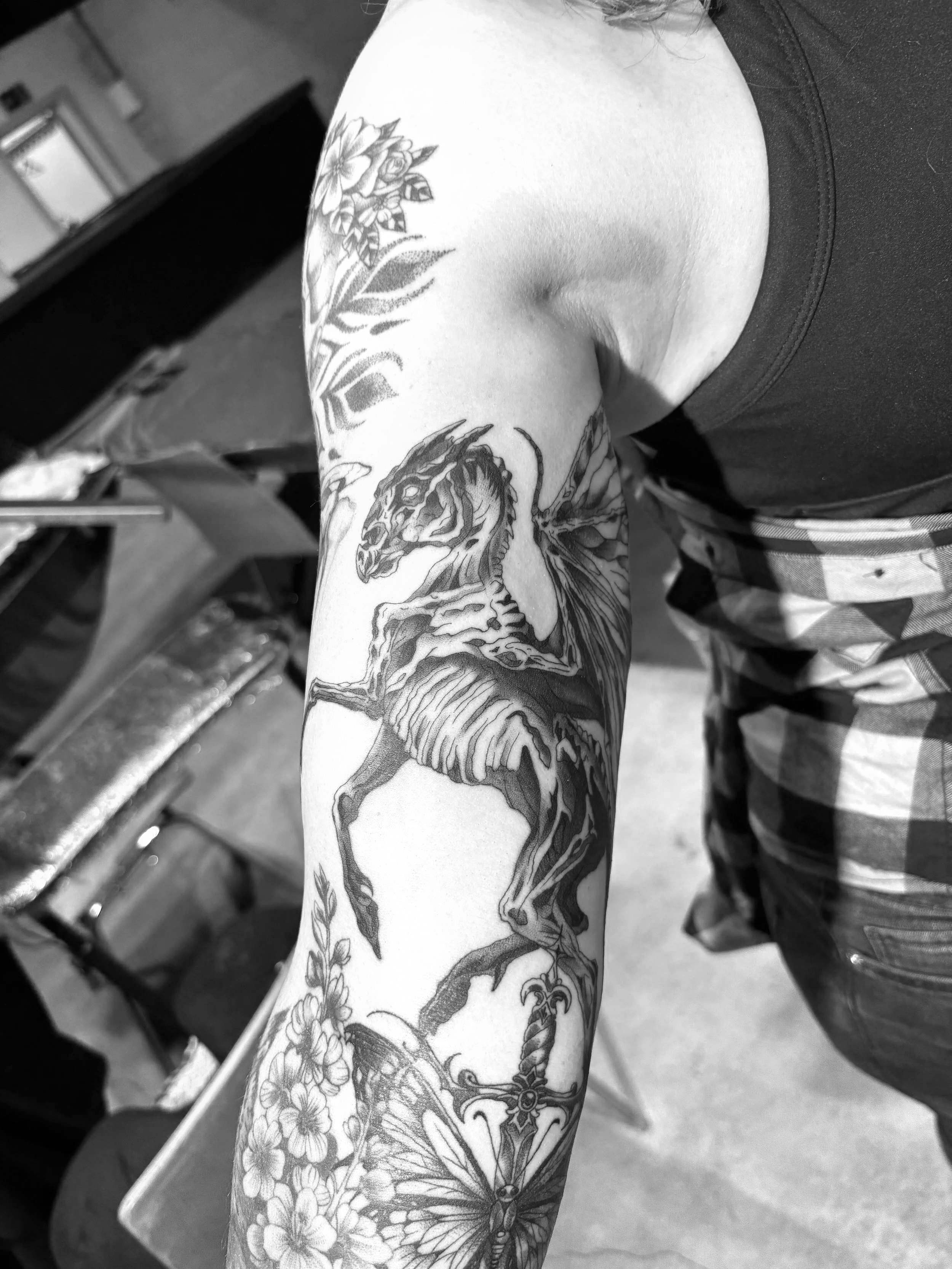 Schwarz-Weiß-Foto eines Arms mit einem detailreichen Tattoo eines springenden Fabelwesens, umgeben von Blumen und Schmetterlingen.