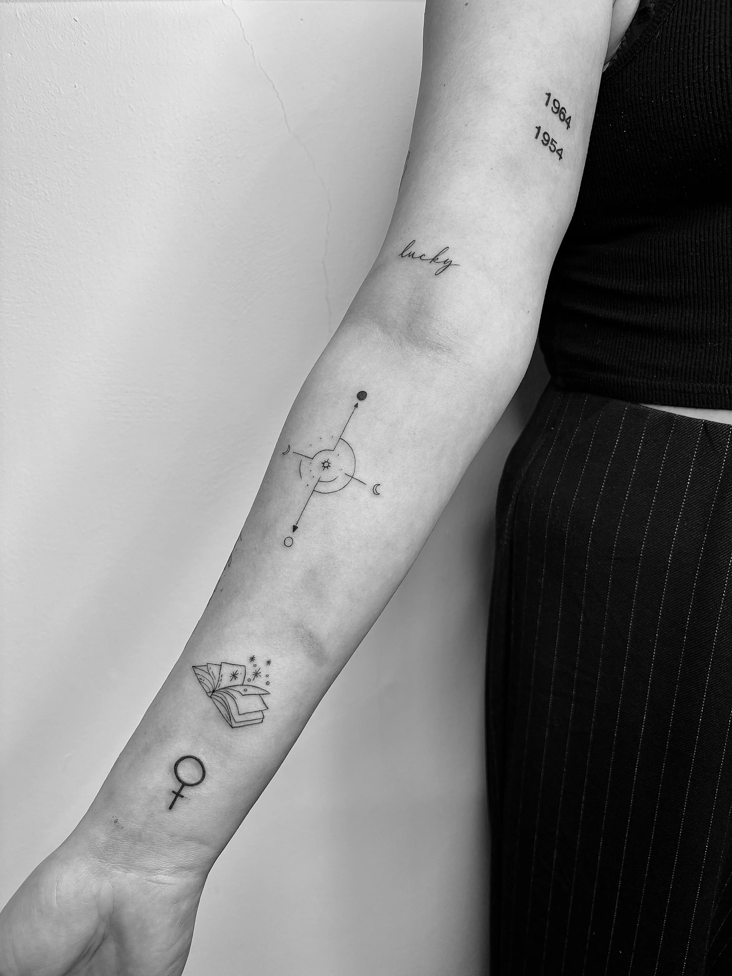 Schwarz-weiße Abbildung eines Arms mit mehreren Tattoos, darunter Zahlen, Wörter wie 'lucky', geometrische Formen, ein Buch und symbolische Darstellungen.