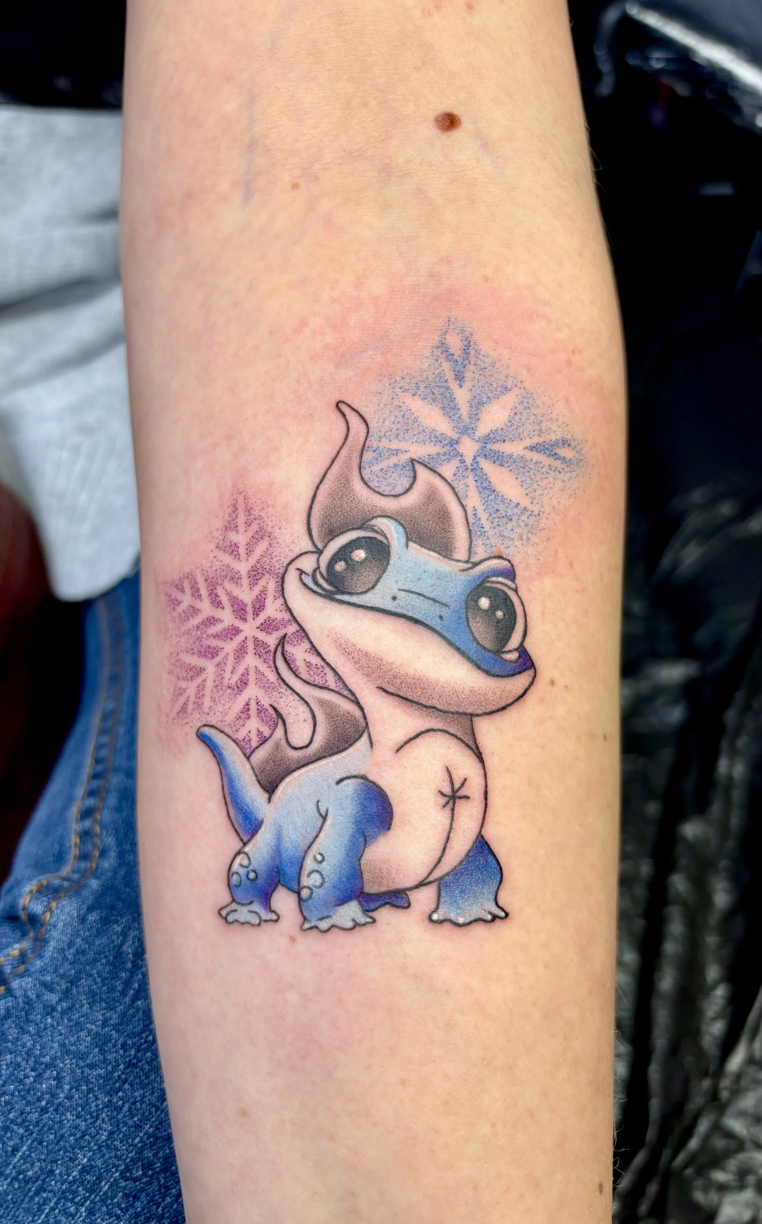 Bunte Tattoo eines kleinen, blauen Dinosauriers auf dem Arm mit Schneeflocken im Hintergrund.