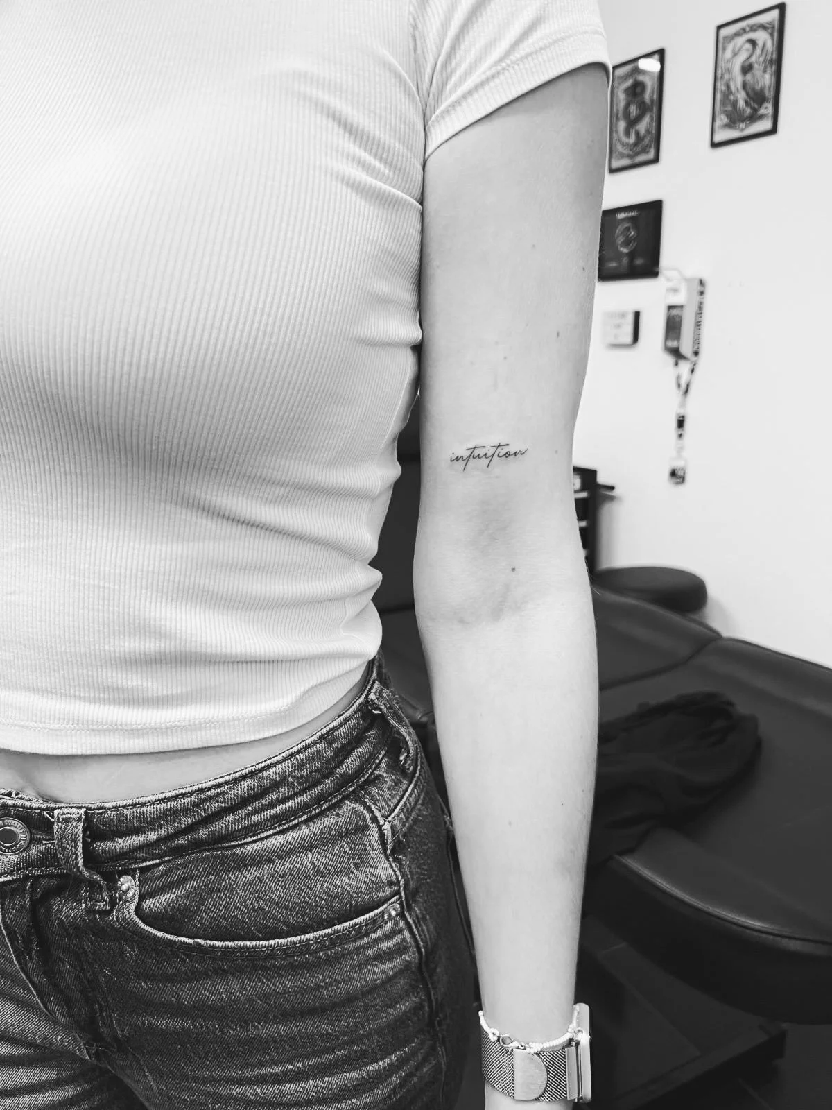 Schwarzer und weißer Tattoo-Schriftzug 'intuition' auf einem Unterarm, teilweise sichtbares weißes T-Shirt, Jeans, Armband, Uhr, Tattoo-Studio im Hintergrund.