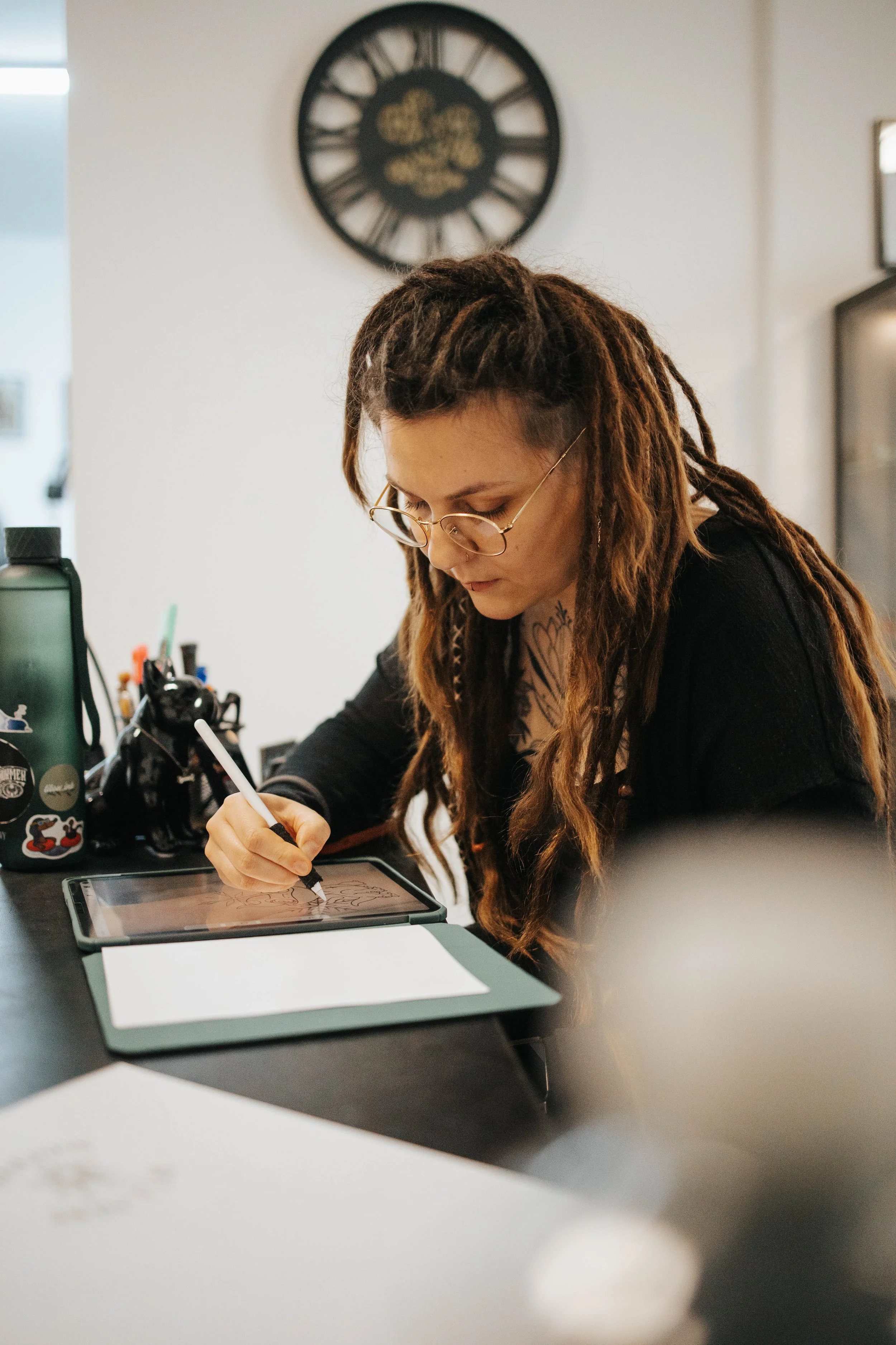 Eine junge Frau mit Dreadlocks und Brille zeichnet mit einem Grafikstift auf einem Grafiktablet. Es befindet sich eine Wasserflasche mit Stickern auf dem Tisch vor ihr.