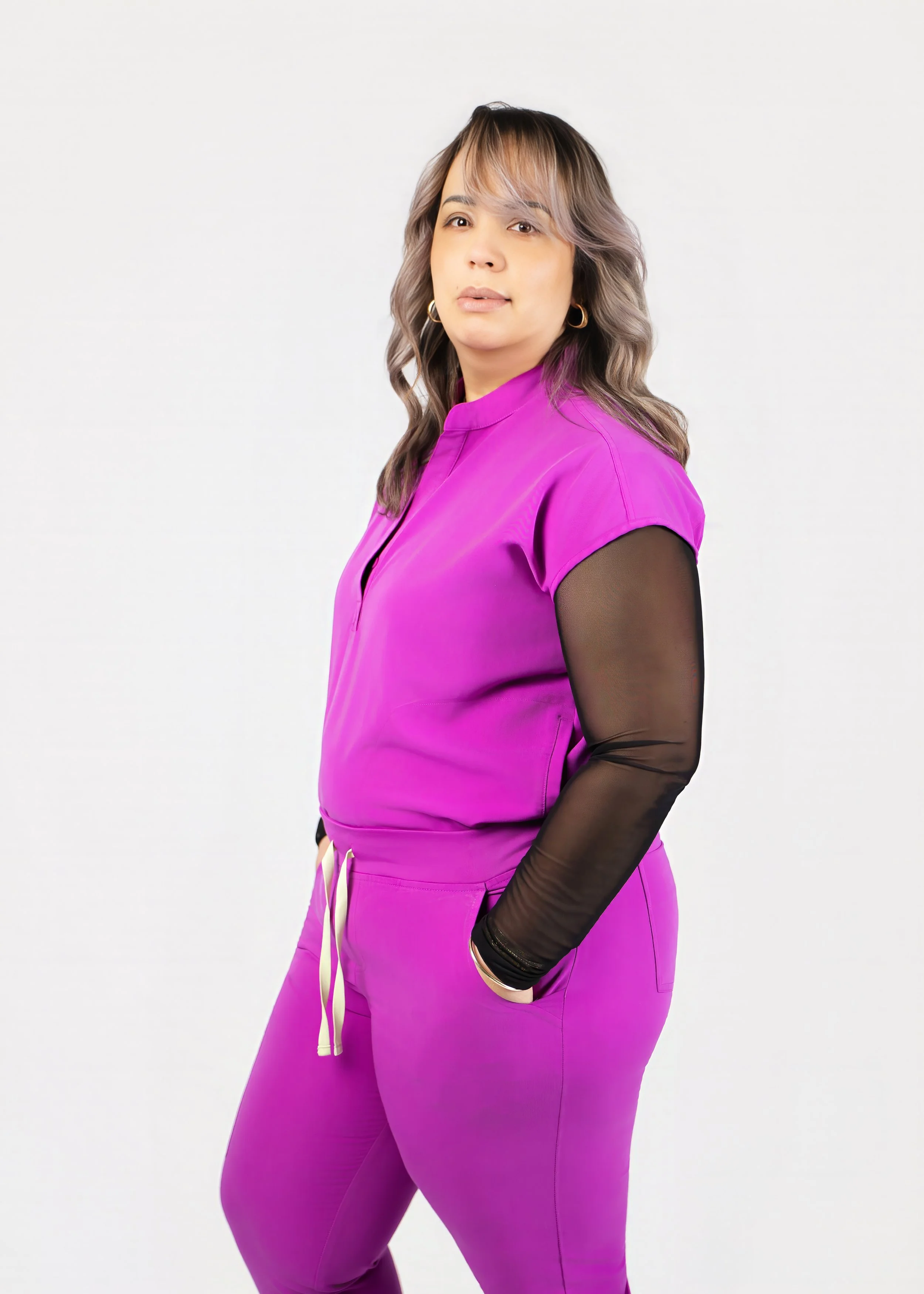 Woman with wavy, light brown hair in a magenta jumpsuit with black sheer long sleeves, standing against a white background.