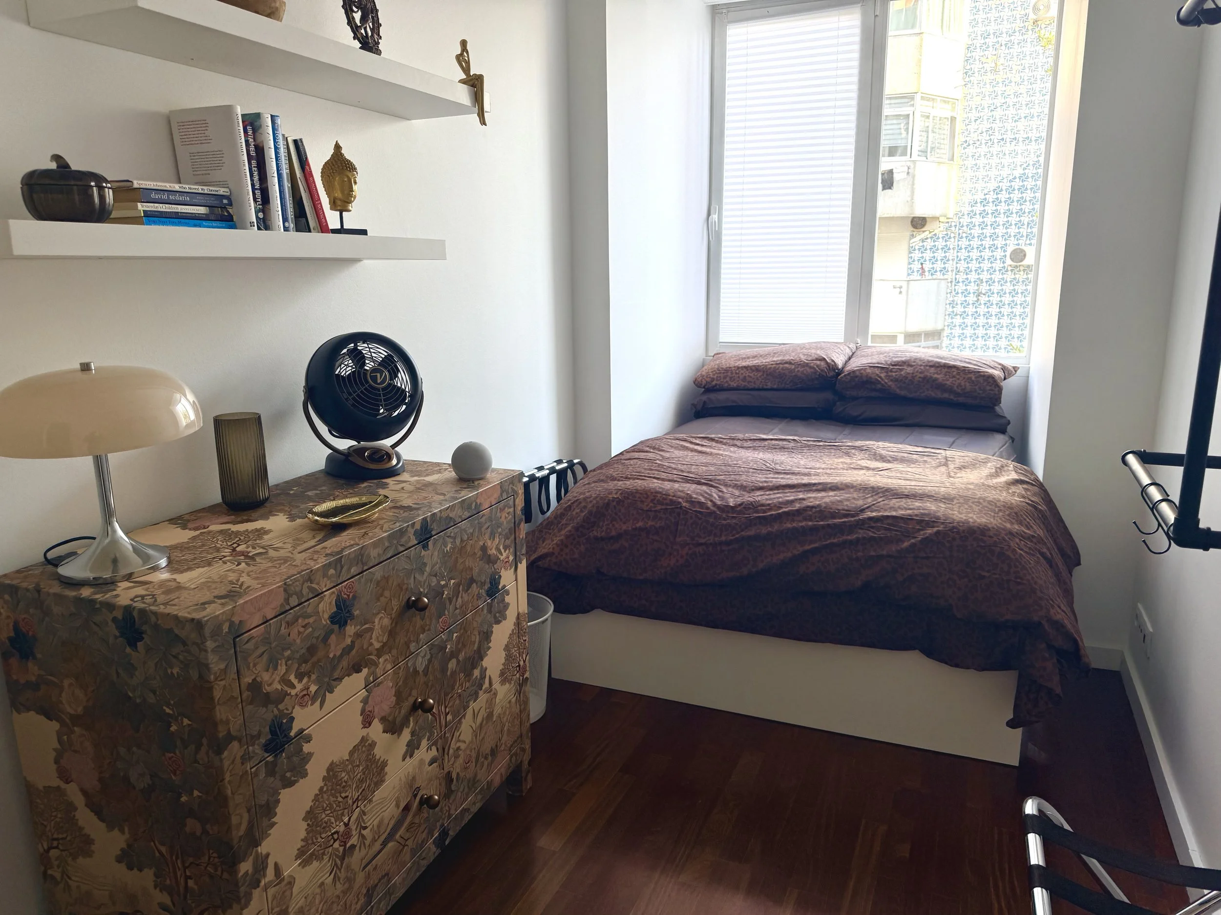 A bedroom with a bed near a large window, a vintage floral dresser, various decorative items on the wall shelves, and an exercise bar in the foreground.