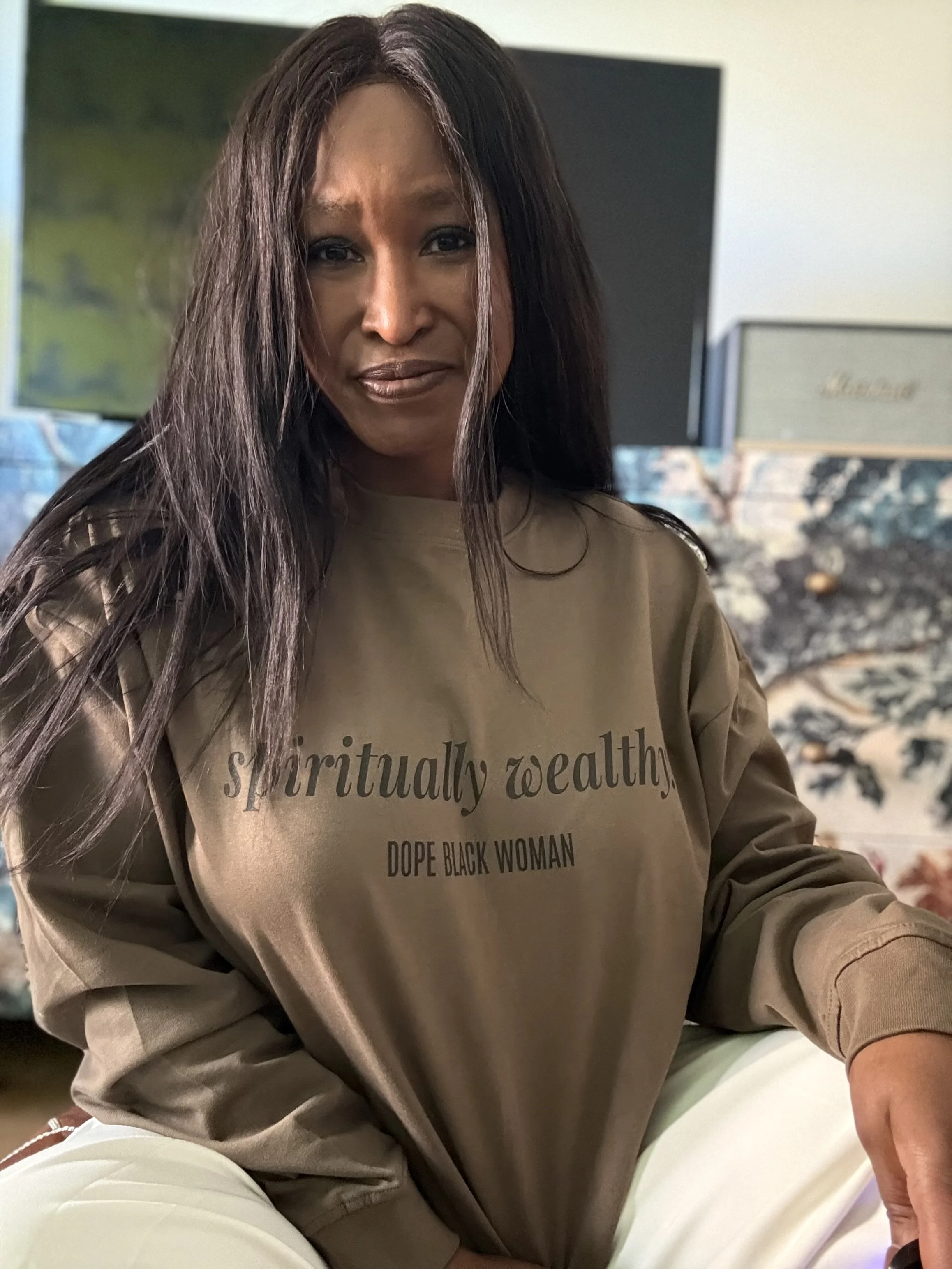 Close-up of a woman with long dark hair wearing a khaki sweatshirt with the words 'spiritually wealthy, dope black woman' printed on it, sitting indoors with a blurred background.