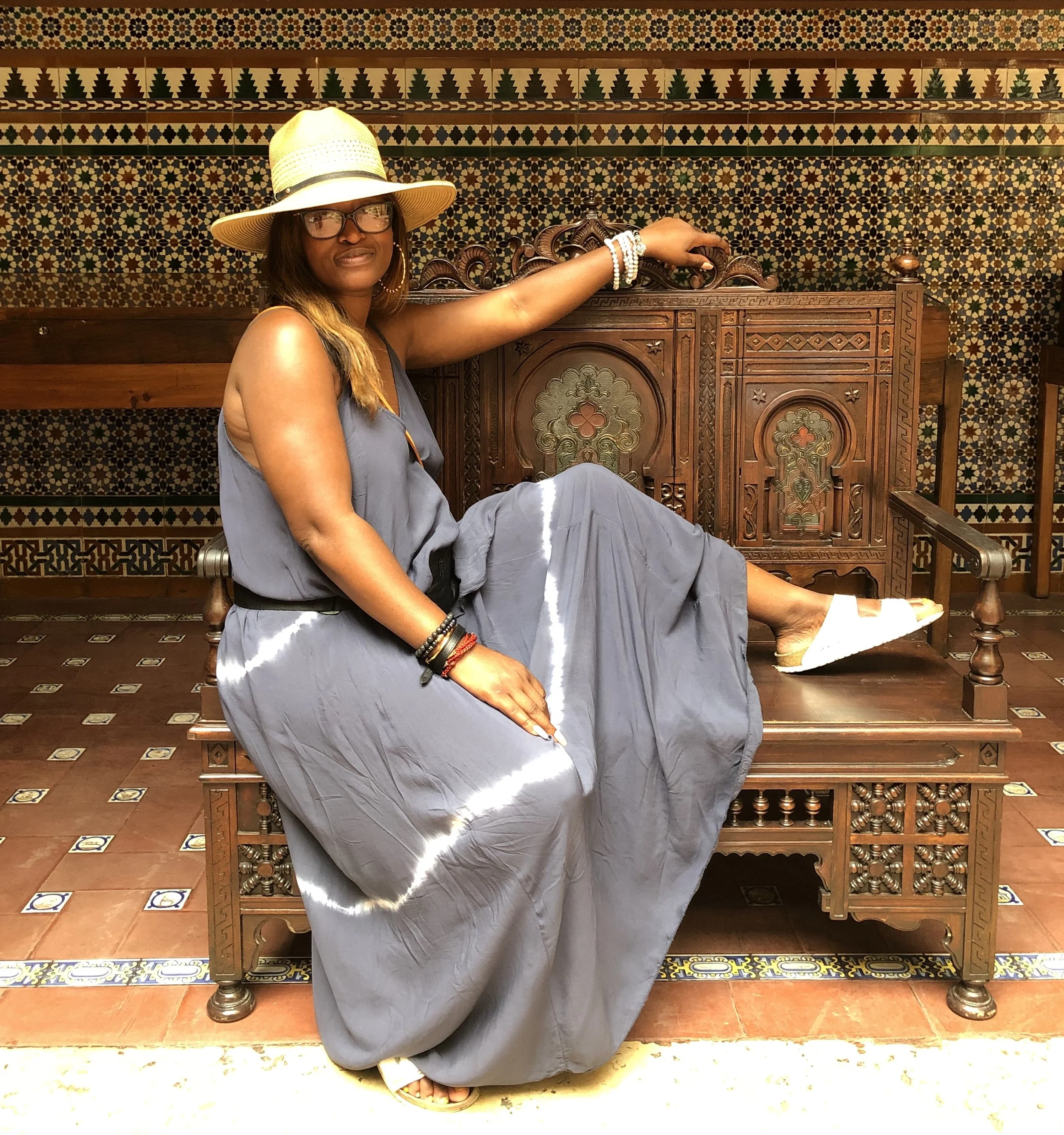 A woman sitting on a wooden bench in a room with Spanish-style tile walls, wearing a large sun hat, glasses, jewelry, and a flowing gray dress, with her foot extended and resting on the bench.