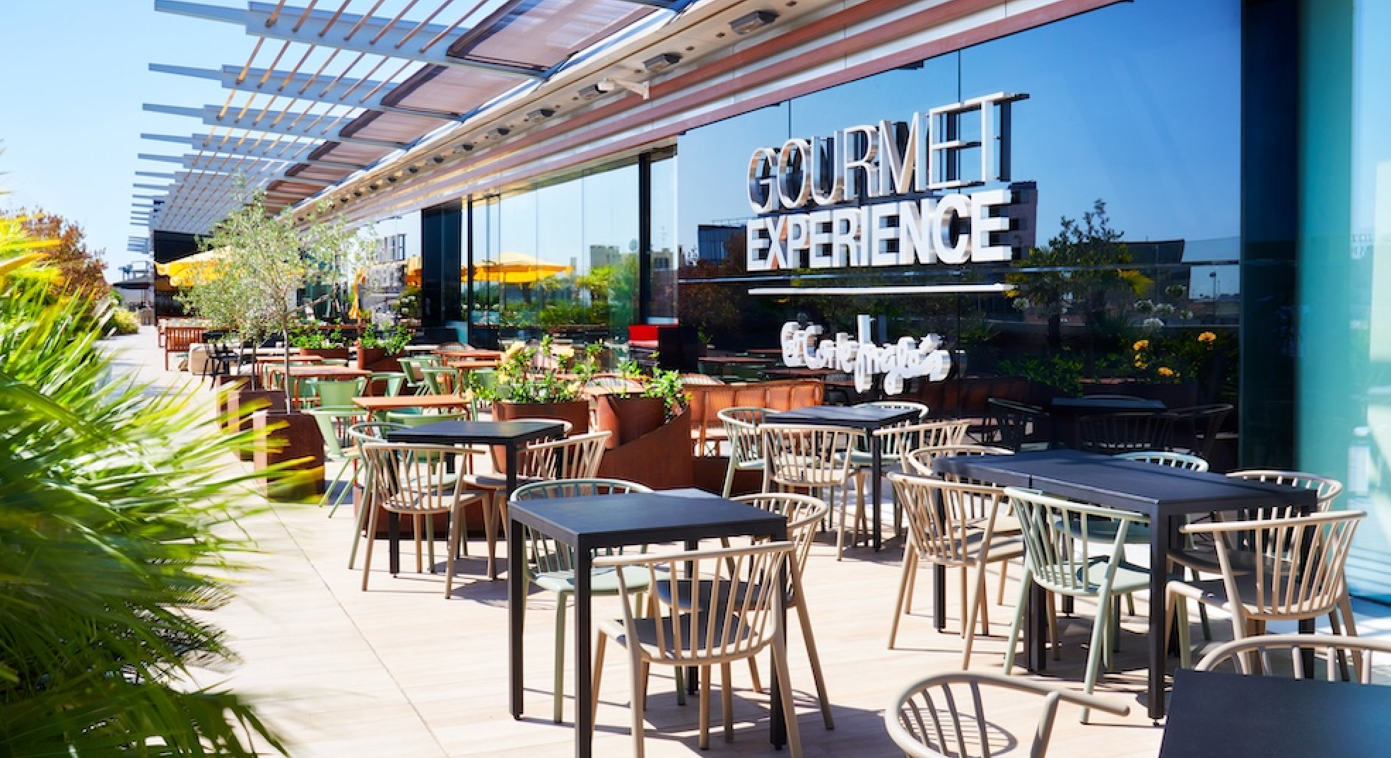 Outdoor patio seating area with tables and chairs in front of a building with large glass windows that have the sign 'GOURMET EXPERIENCE' on them. There are plants and yellow umbrellas visible in the background.