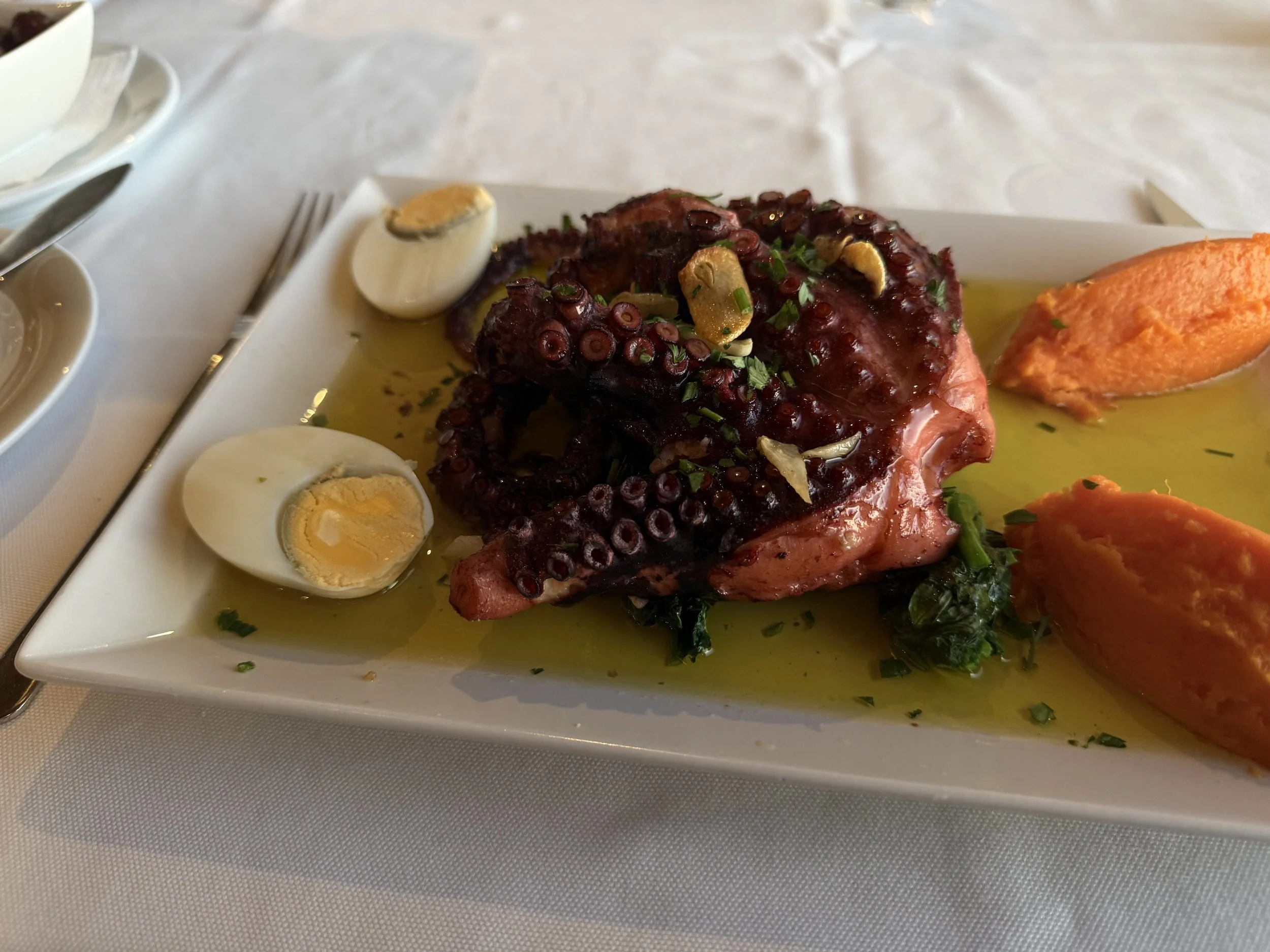 A plate of cooked octopus garnished with chopped herbs, garlic slices, and served with halved boiled eggs and a dollop of orange-colored sauce or puree.