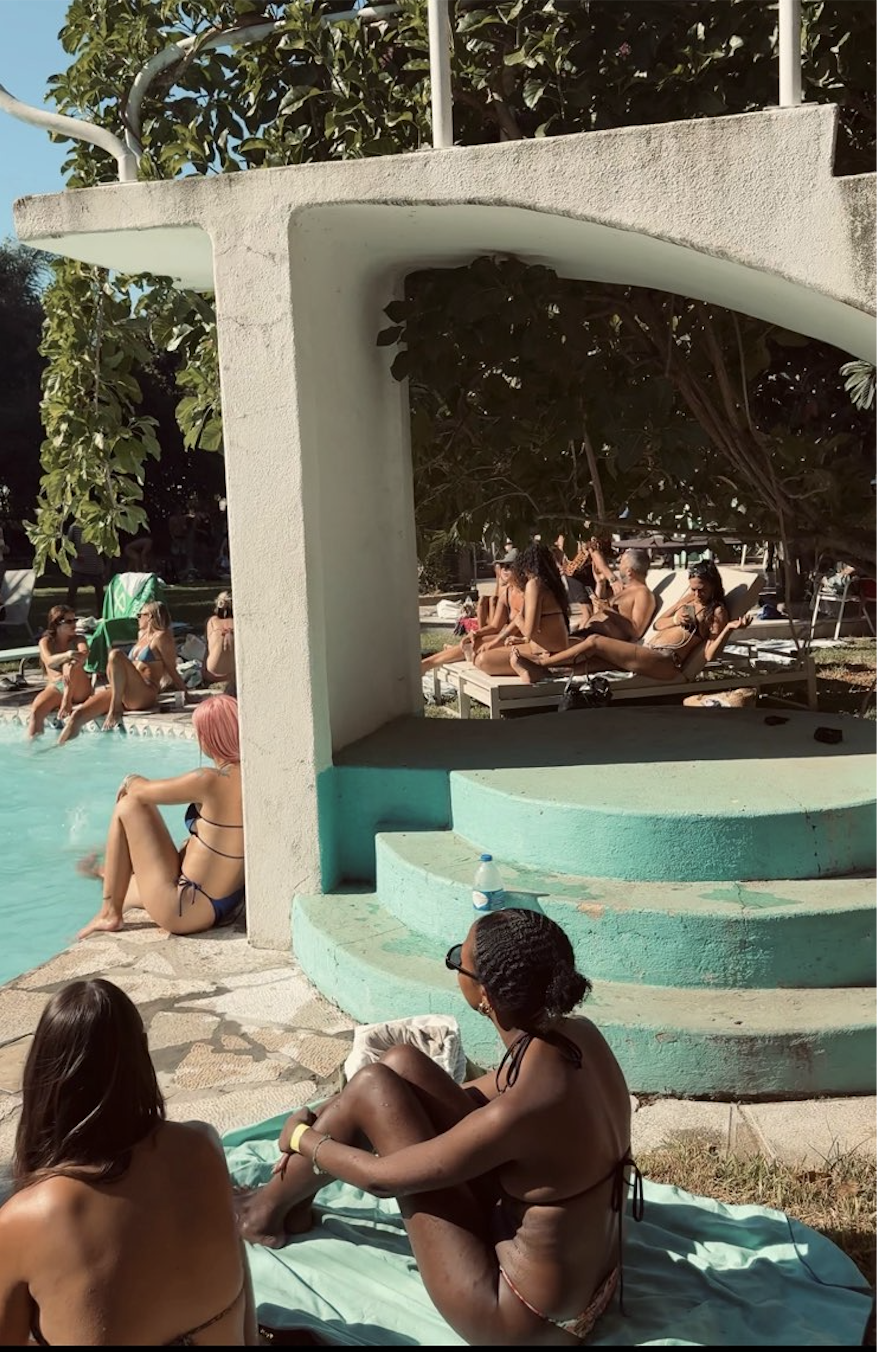 People relaxing and socializing poolside at a summer outdoor pool, with some sitting on lounge chairs and others in the pool, under a shaded area with trees in the background.