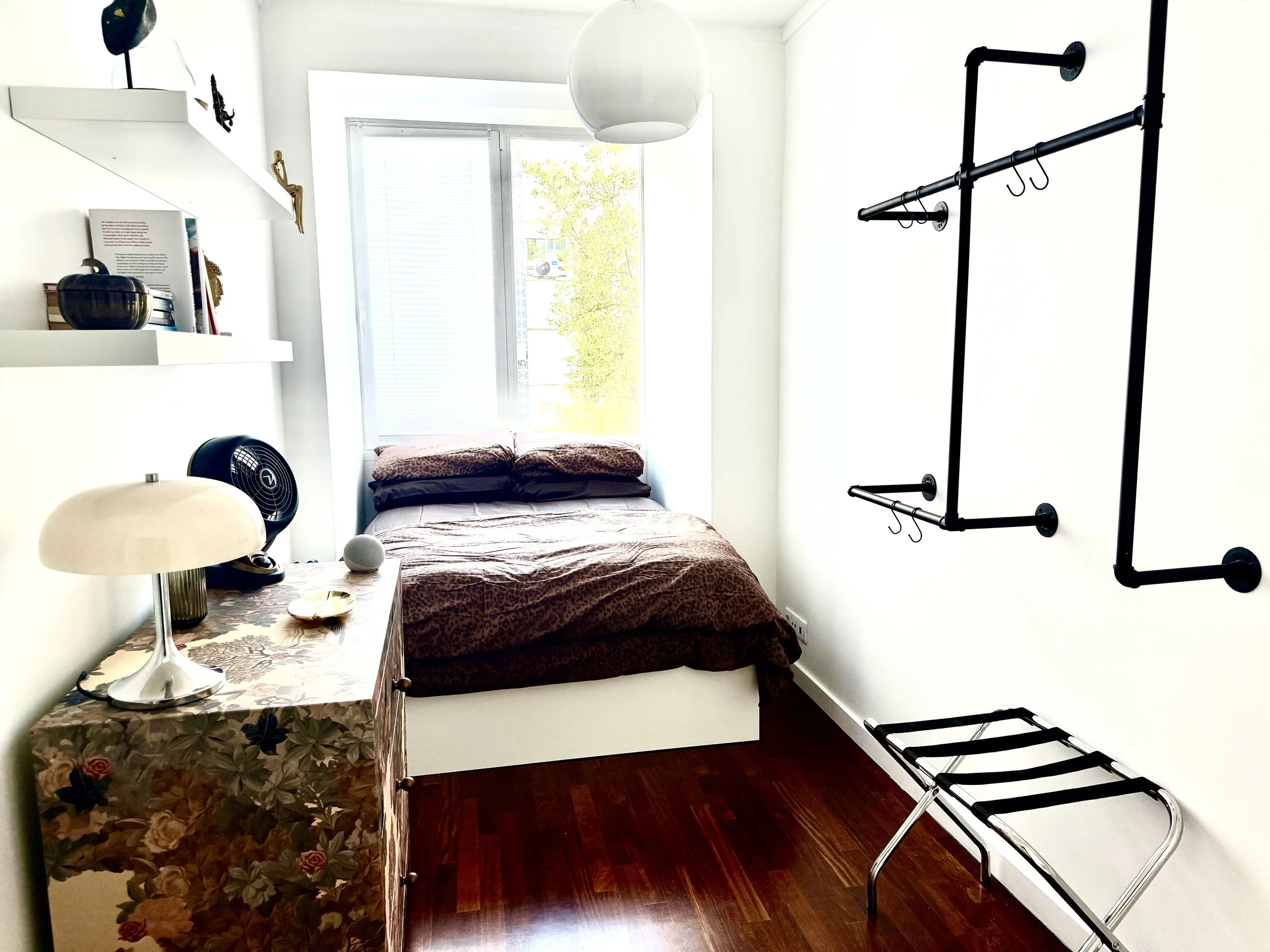 A small bedroom with a bed covered in a leopard print blanket placed under a large window with a view of trees outside. To the left, a bedside table with a table lamp, a decorative bowl, and some books. Above the bed, a black wall-mounted exercise pull-up bar. To the right, a black clothing rack and a chrome luggage rack. The room has white walls and dark wooden flooring.