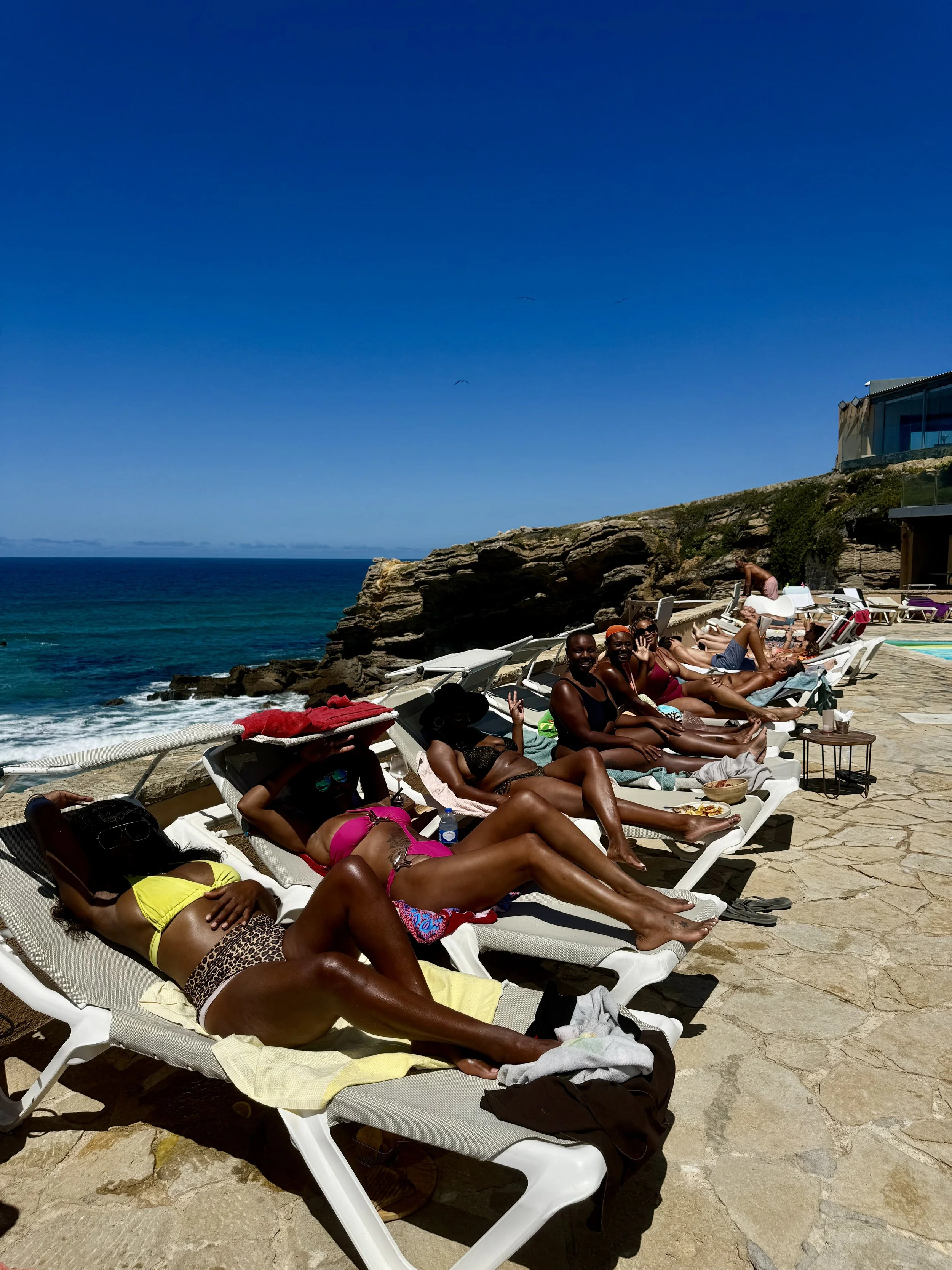 People sunbathing and relaxing on lounge chairs by the beach under a clear blue sky with ocean in the background.