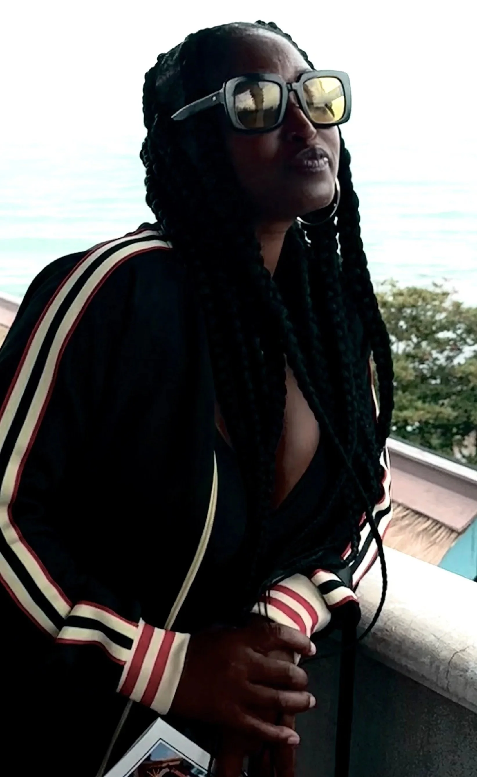 Woman with dark skin and black braids wearing large sunglasses and a black jacket with red, white, and beige stripes on the sleeves, standing near a body of water with trees in the background.
