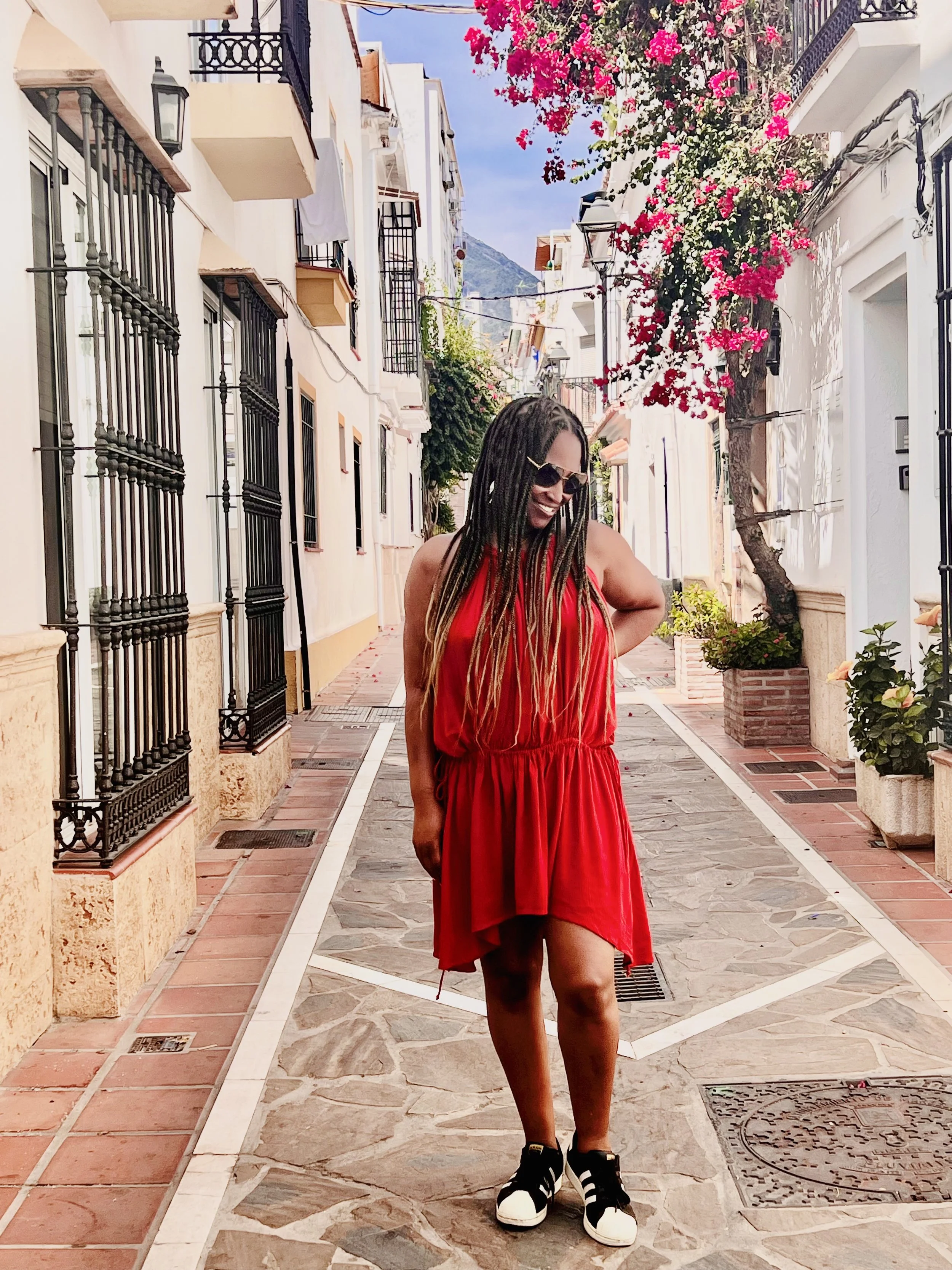 A woman in a red dress and black Adidas sneakers standing on a cobblestone street lined with white buildings, black iron balconies, and pink flowering trees under a blue sky.