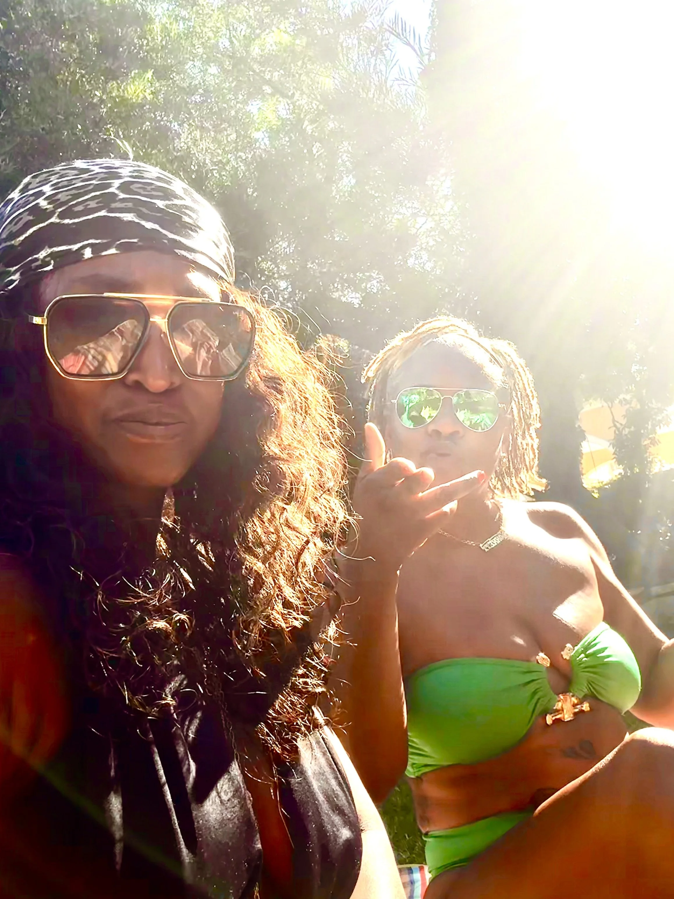 Two women outdoors in bright sunlight, one with curly hair, sunglasses, headscarf, and gray hoodie; the other with curly hair, sunglasses, and a green swimsuit top, gesturing with her hand.