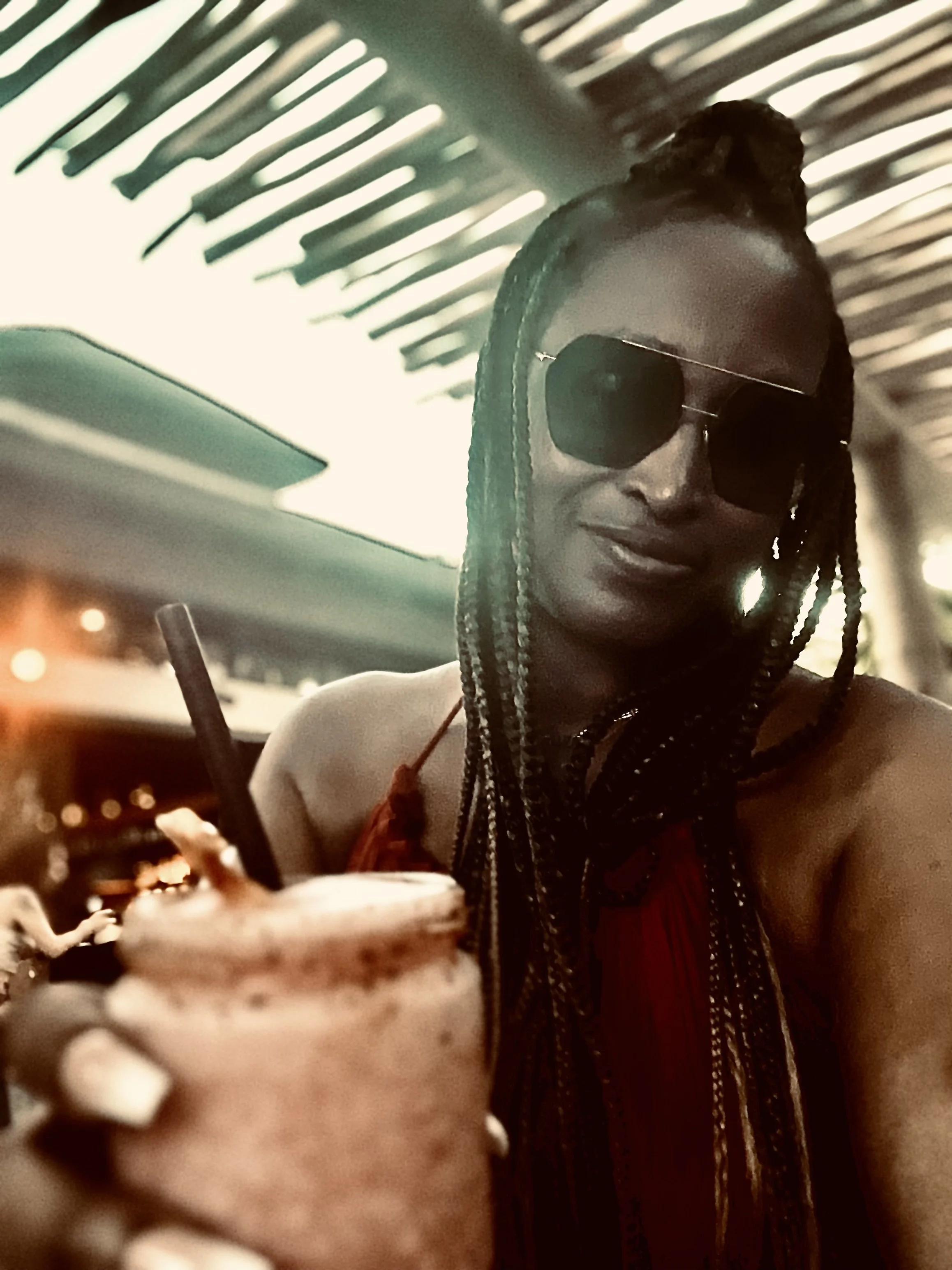 A woman with braided hair, wearing large sunglasses and a red dress, sitting at a table in a restaurant or cafe while having a dessert topped with whipped cream and ice cream, with a drink nearby.