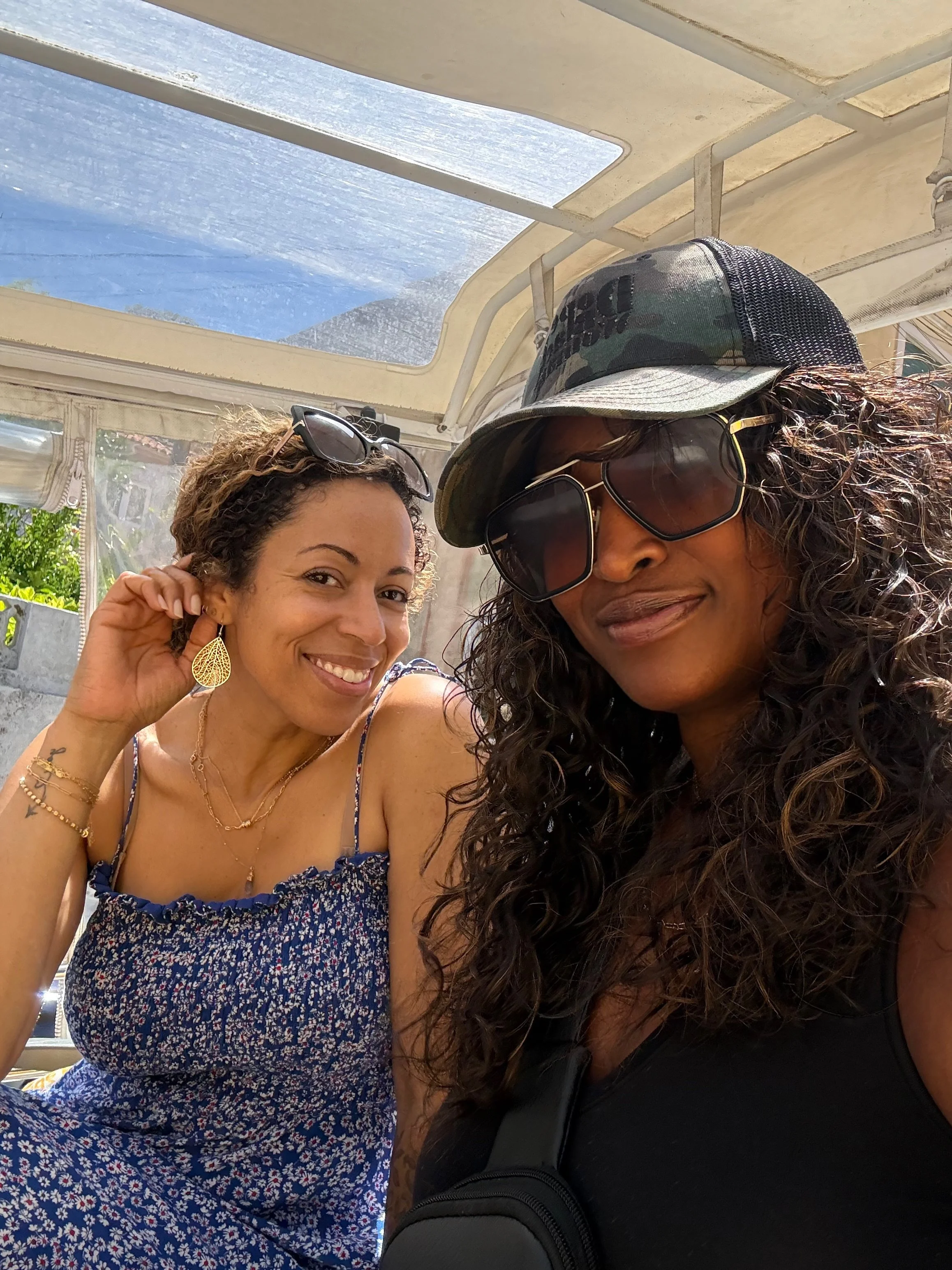 Two women taking a selfie together outdoors. One woman has curly, shoulder-length hair, and is wearing a blue floral dress, gold jewelry, and sunglasses on her head. The other woman has long, curly hair, large sunglasses, a camouflage cap, and a black top. They appear to be smiling and enjoying their time together.
