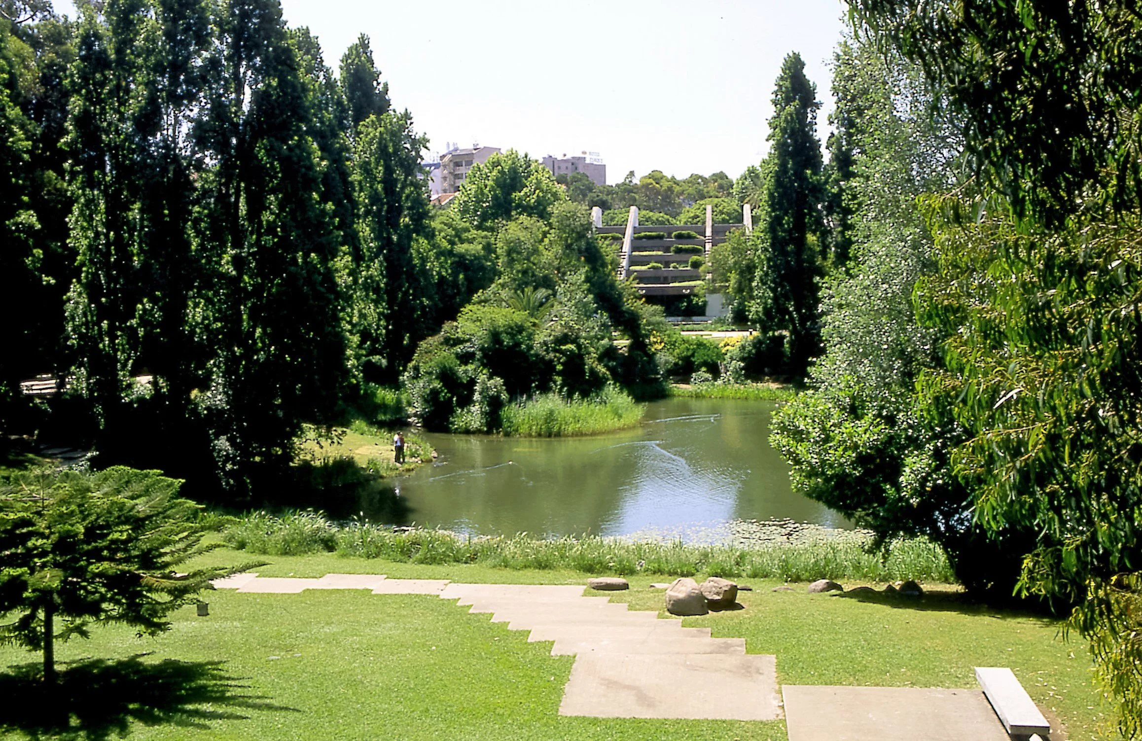 A landscaped park with a small river, lush green trees, and a concrete staircase leading to a raised area with a decorative bridge in the background.