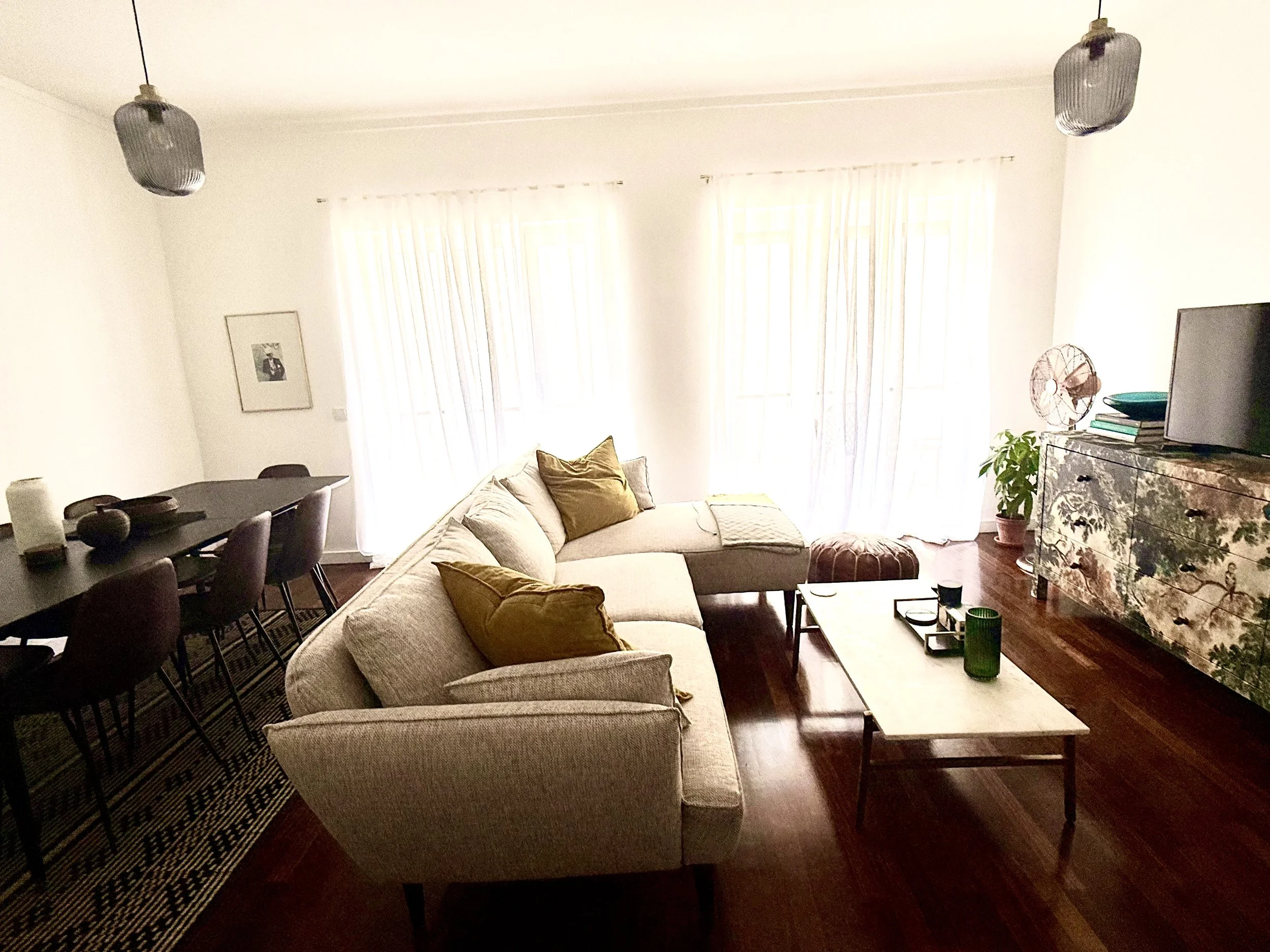 Living room with large windows, beige sectional sofa with brown and yellow pillows, wooden coffee table, patterned rug, dining table with chairs, fireplace with decorative cover, TV, and various plants and decor.