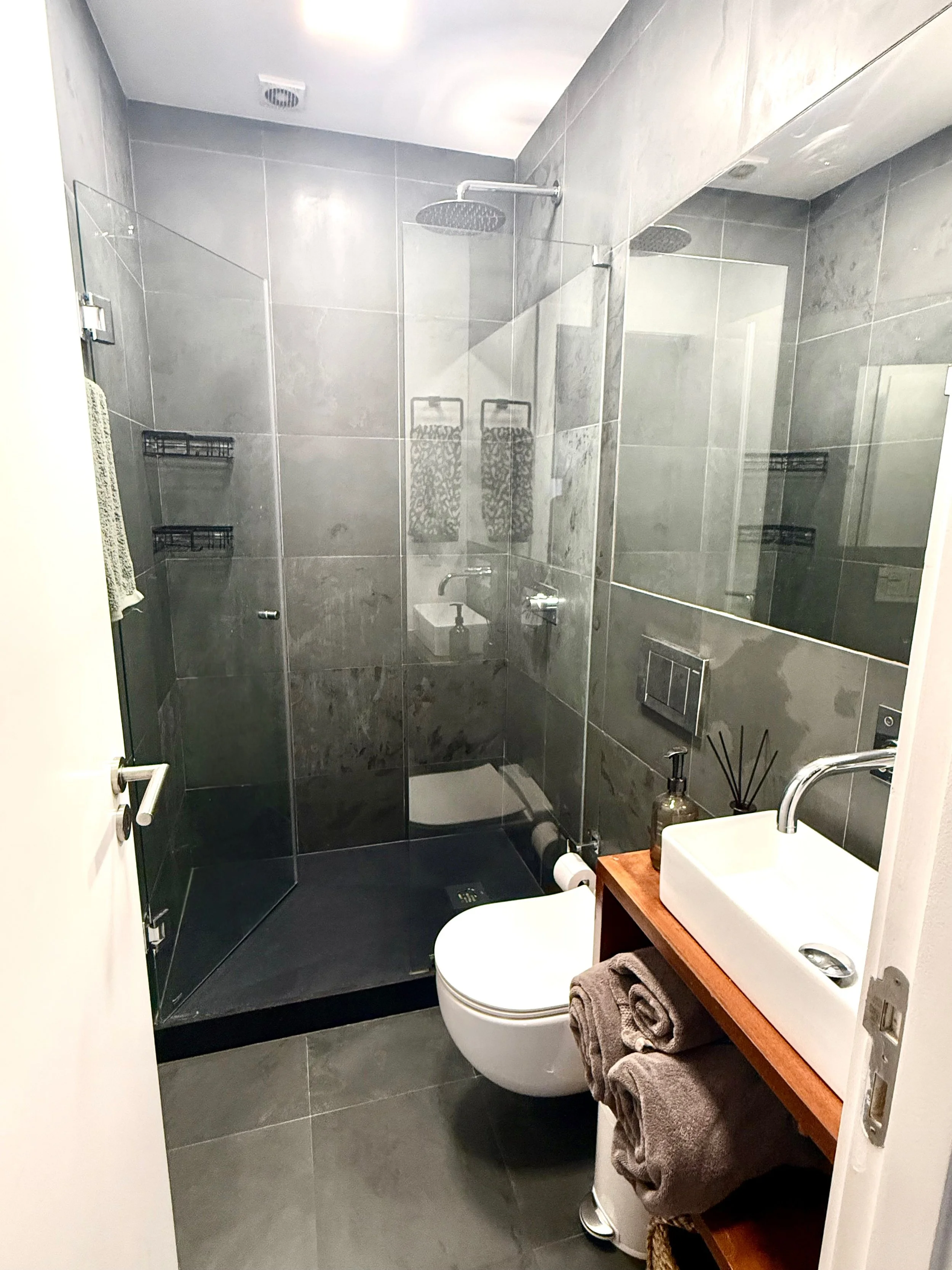 Modern bathroom with dark gray tiled walls and floor, a walk-in shower with glass door, a wall-mounted toilet, a wooden vanity with a rectangular sink, a mirror, and decorative accessories.