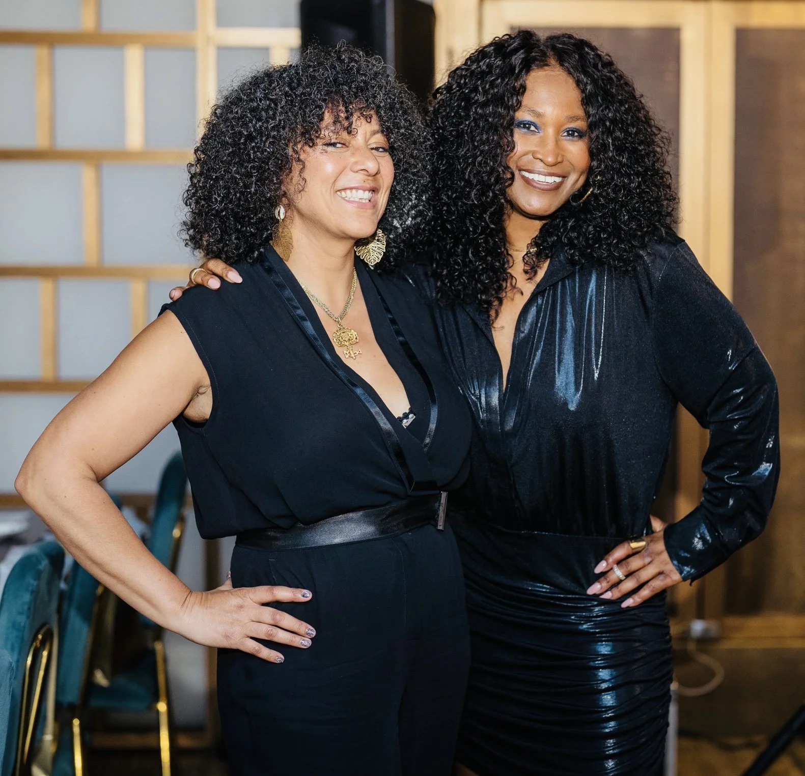 Two women with curly hair and dark clothing, smiling and posing together indoors.