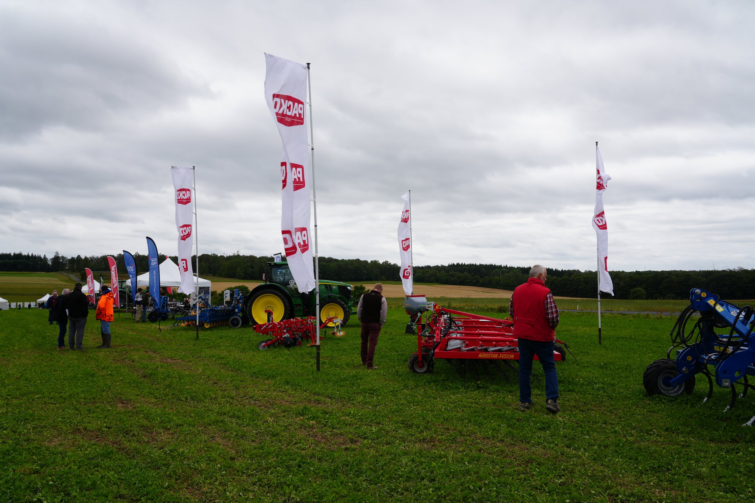 Vente d'équipements agricoles sur un stand en plein air avec plusieurs drapeaux blanc et rouge portant le logo 'PAG' et d'autres équipements agricoles exposés. Plusieurs personnes visitent le stand, sous un ciel nuageux dans un champ avec des arbres à l'arrière-plan.